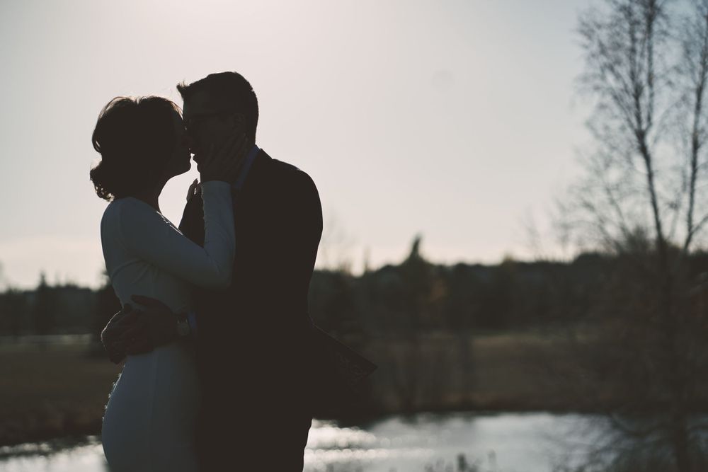 A silhouetted couple embraces by a lake at sunset, creating a romantic backlit scene against a misty sky.