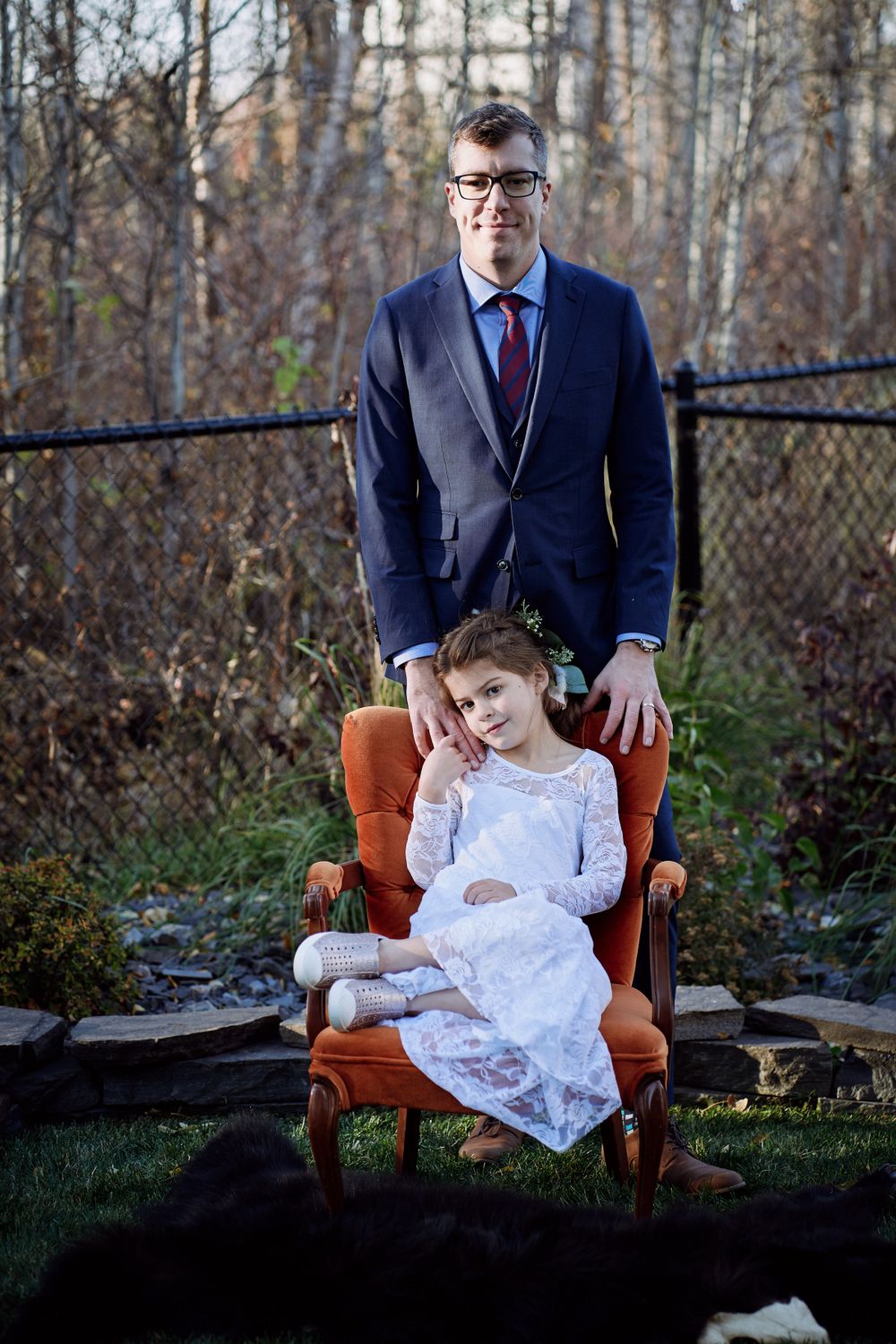 A formal portrait featuring a vintage orange armchair outdoors against a wooded backdrop in autumn.