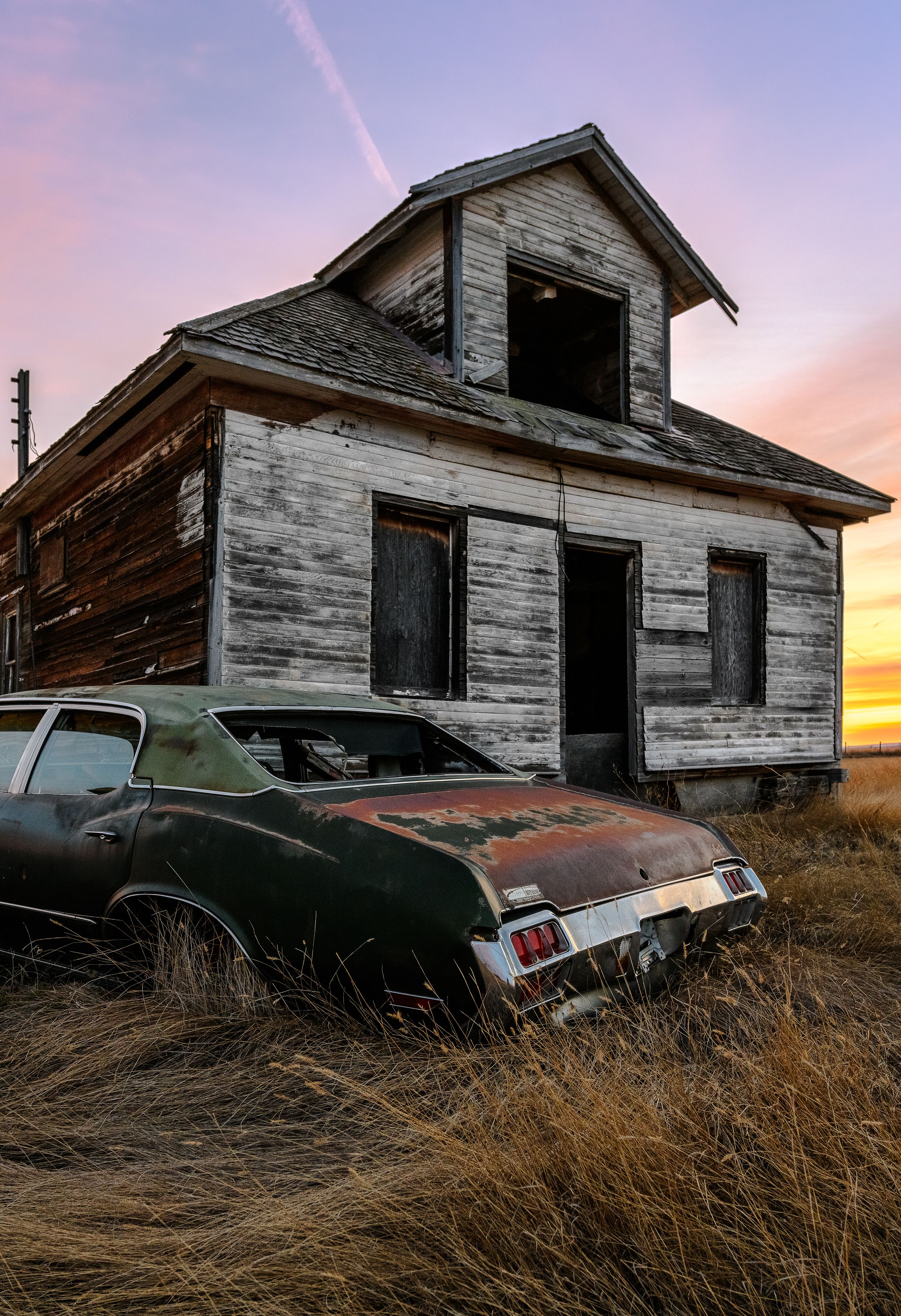 Abandoned wooden house with a vintage rusted car against a dramatic sunset sky.