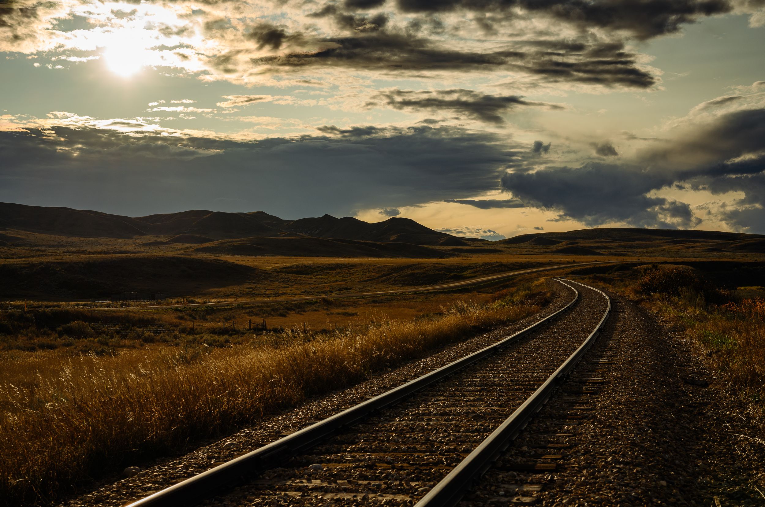 Railroad tracks curve into the distance under a dramatic sunset with mountains on the horizon.