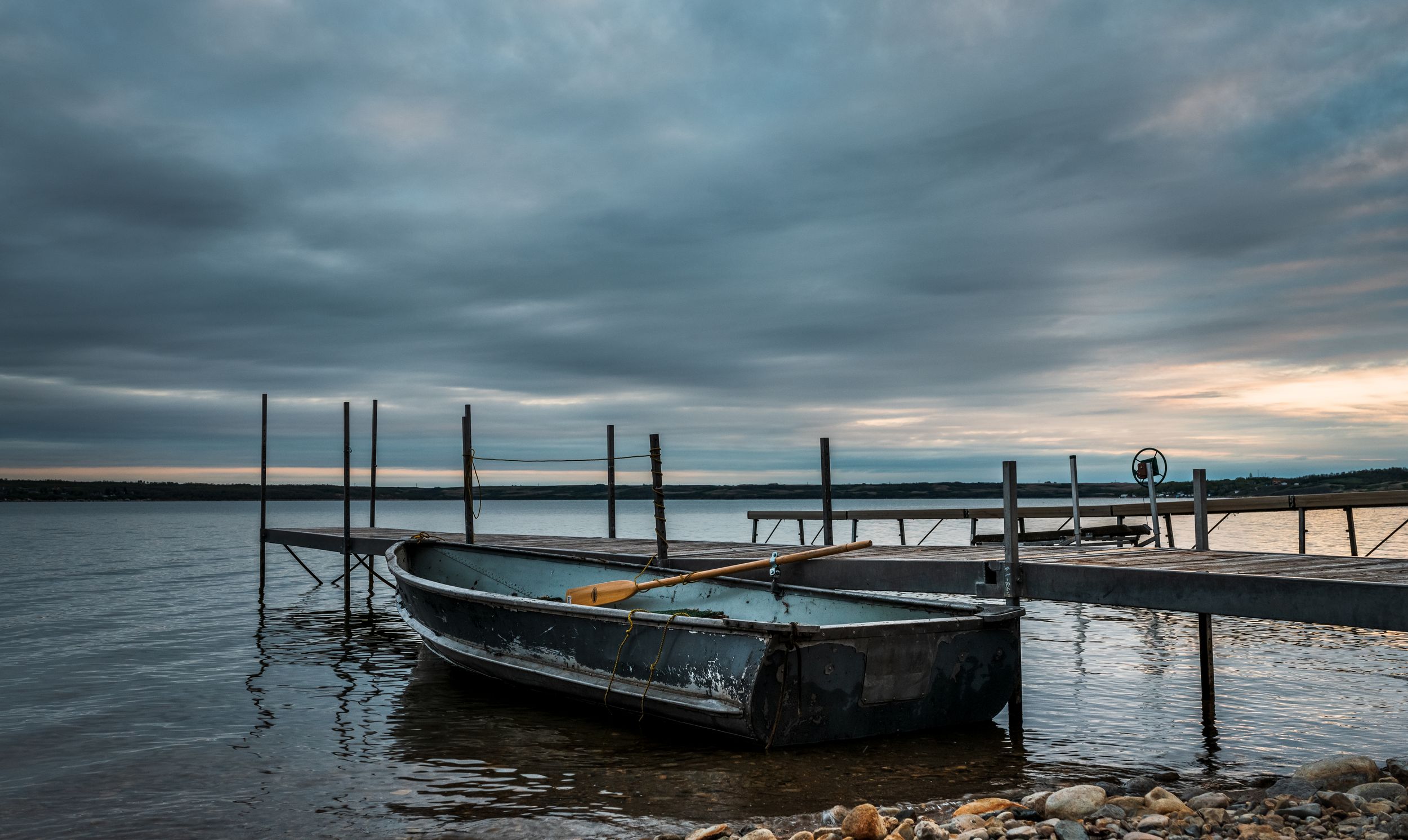 A weathered boat tied to a wooden dock on a cloudy lake during dramatic sunset with moody skies.