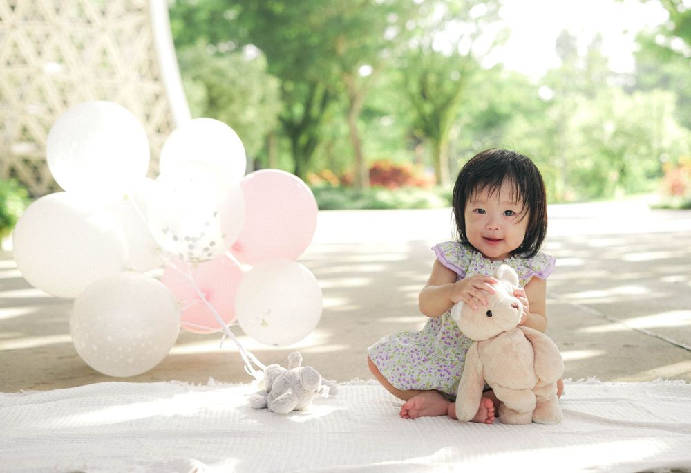Toddler sitting with stuffed animal and white balloons in outdoor garden setting.