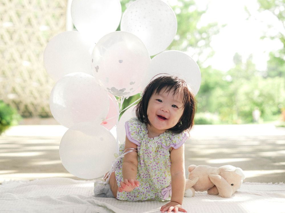 Happy child sitting with white and pink balloons and plush toy in garden.