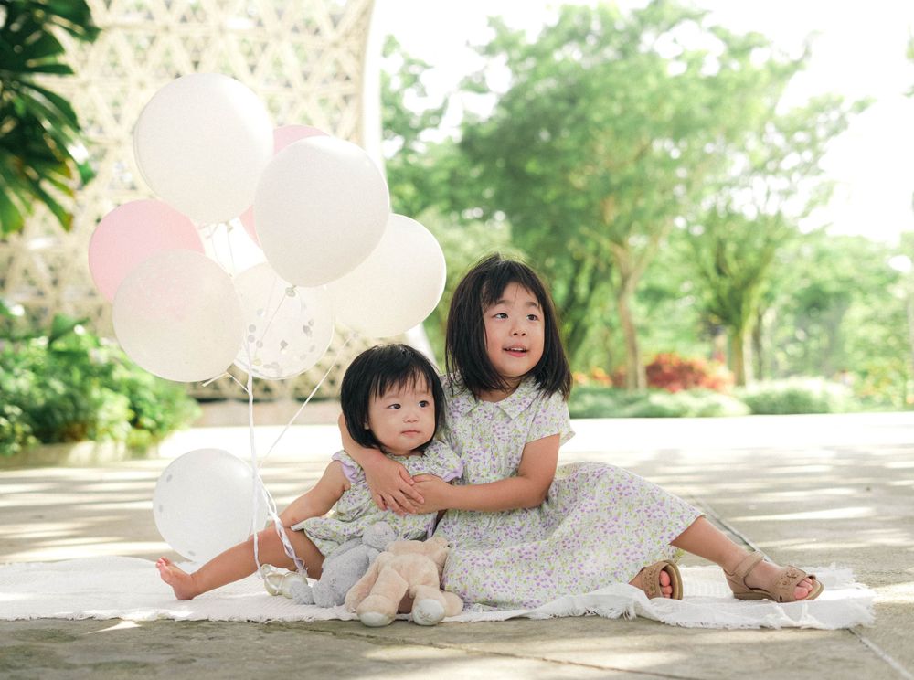 Two children in matching dresses sitting together with white balloons in garden setting.