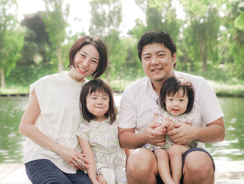 A happy family of four poses together outdoors near a lake on a sunny day.