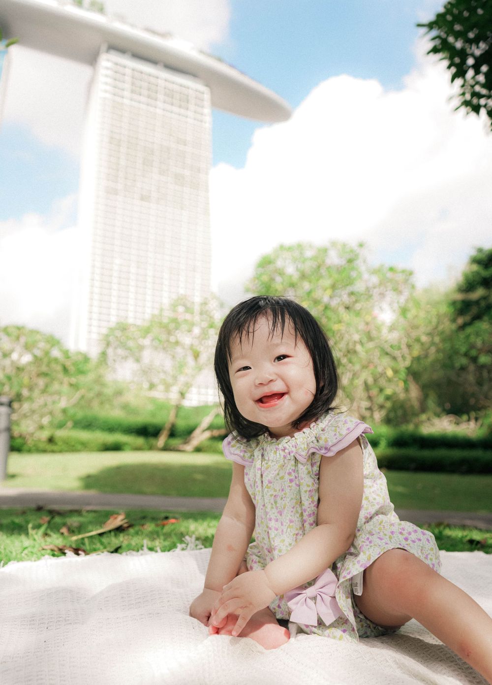A young child in a floral dress sits outside with the Marina Bay Sands building visible in the background.