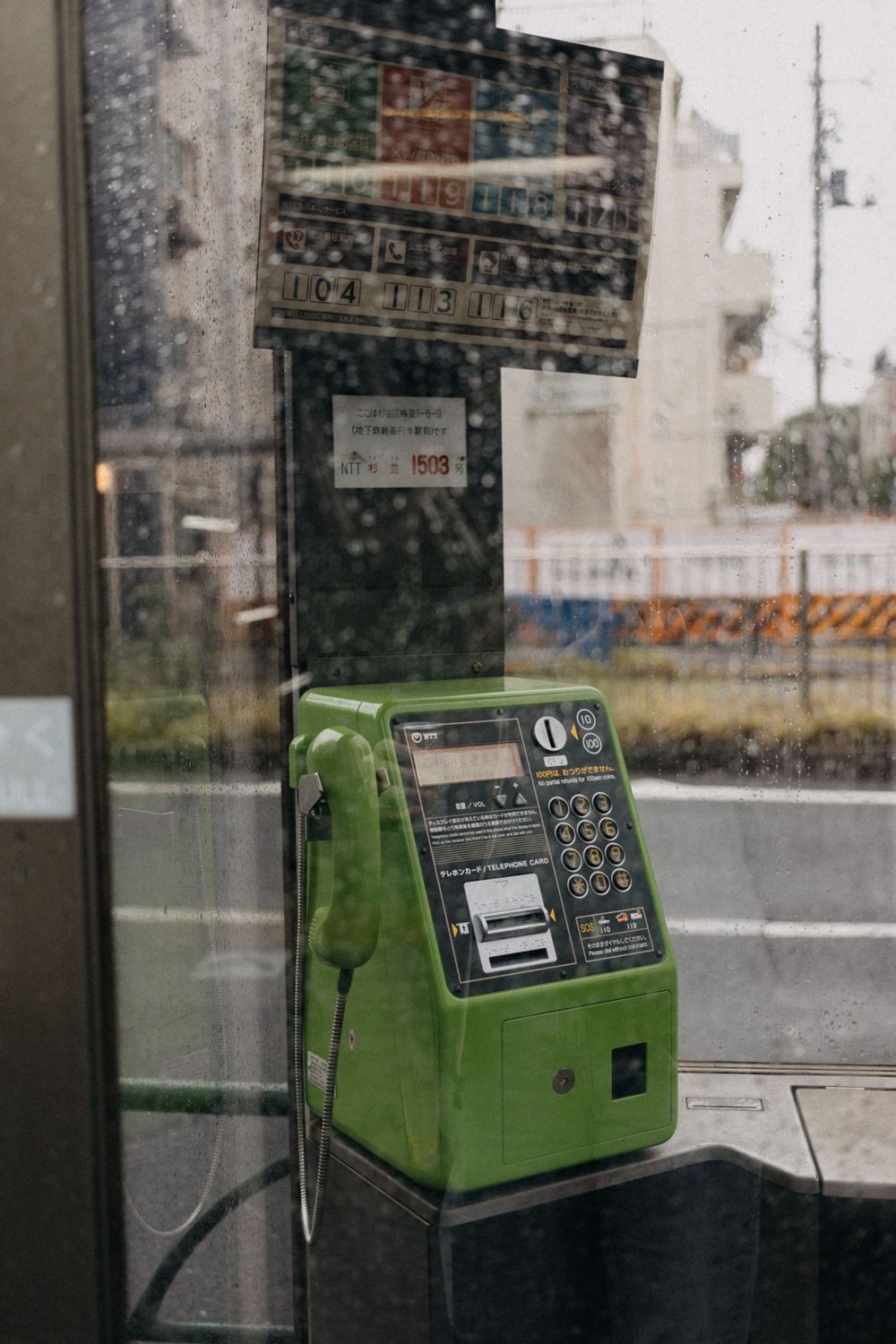 A vintage green public payphone in a glass booth on a rainy Japanese street.