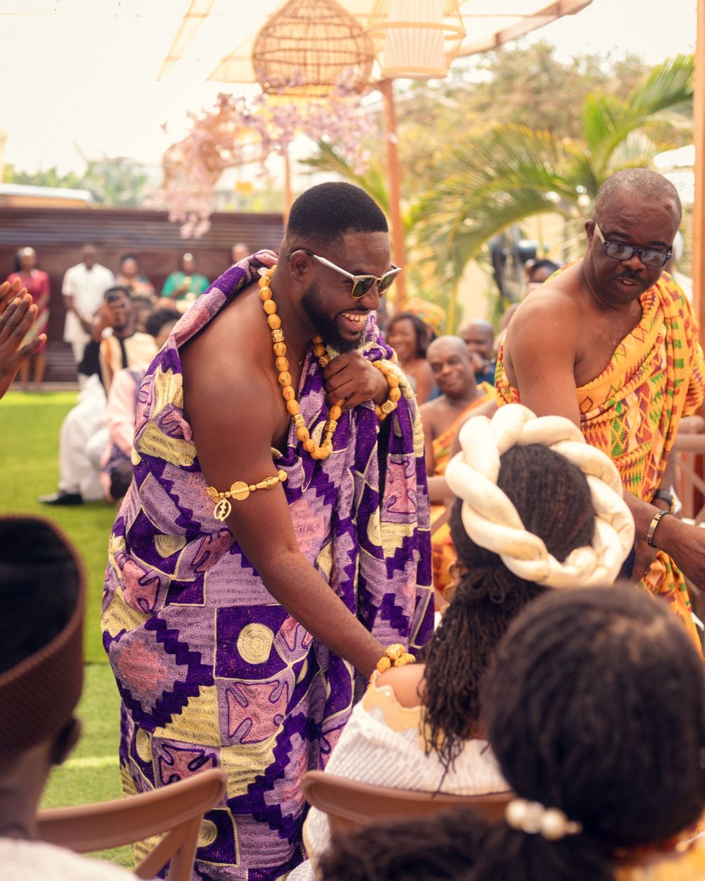 A celebration scene with people in purple traditional African attire at an outdoor event.
