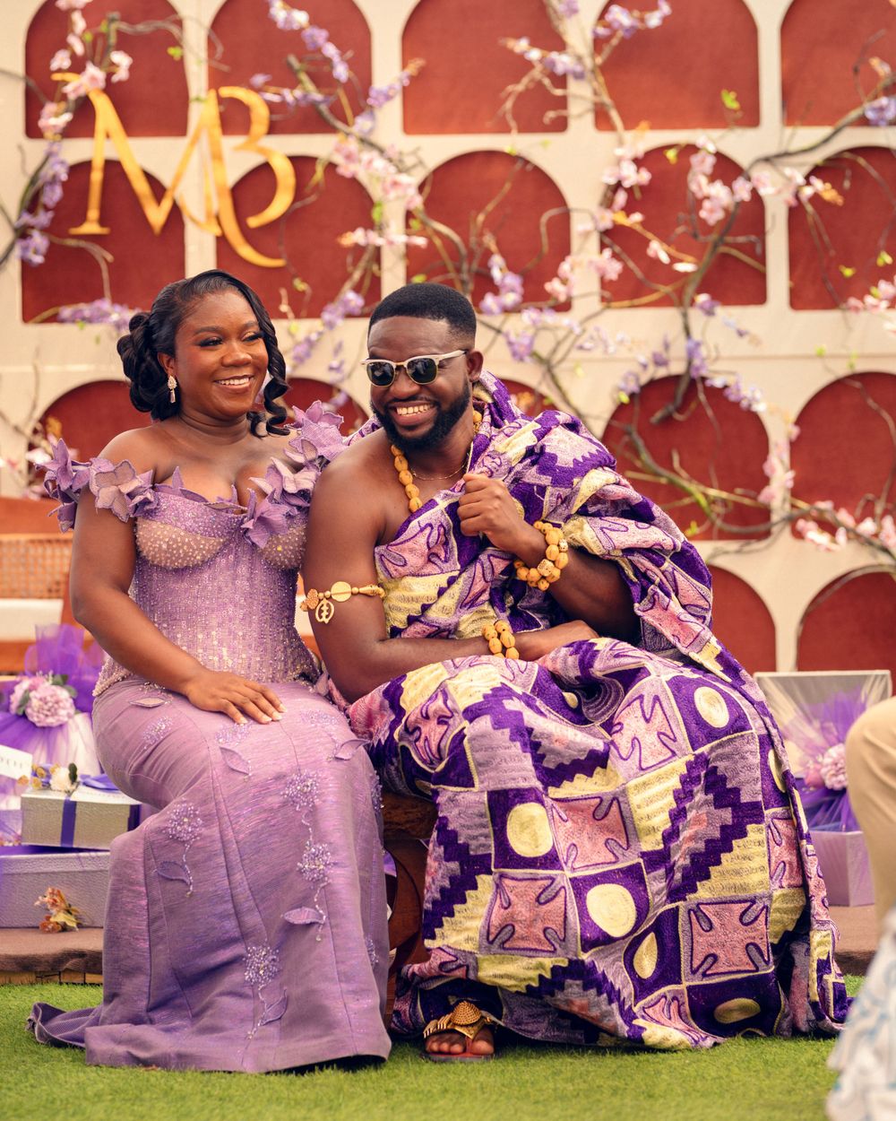 Couple in matching purple formal wear pose together against decorative red wall backdrop.