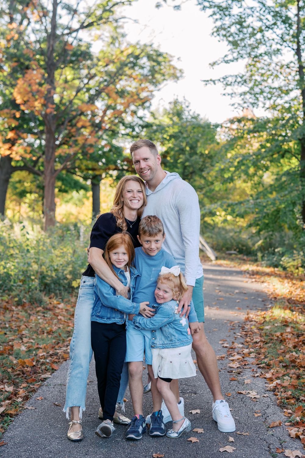 A family hugs close together while standing on an autumn trail surrounded by trees.