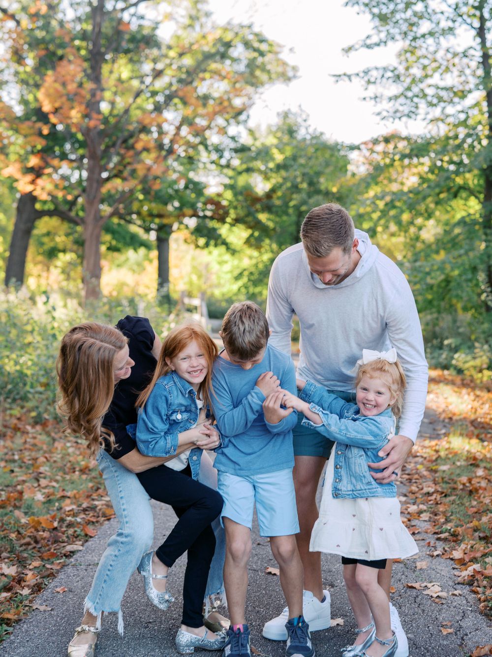 Family plays and laughs together on a tree-lined path covered in autumn leaves.
