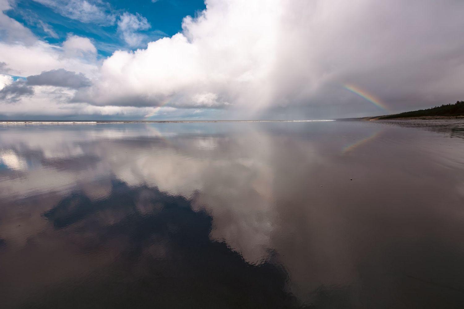 A stunning rainbow appears over a mirror-like beach where clouds reflect perfectly in the wet sand during low tide on Haida Gwaii.