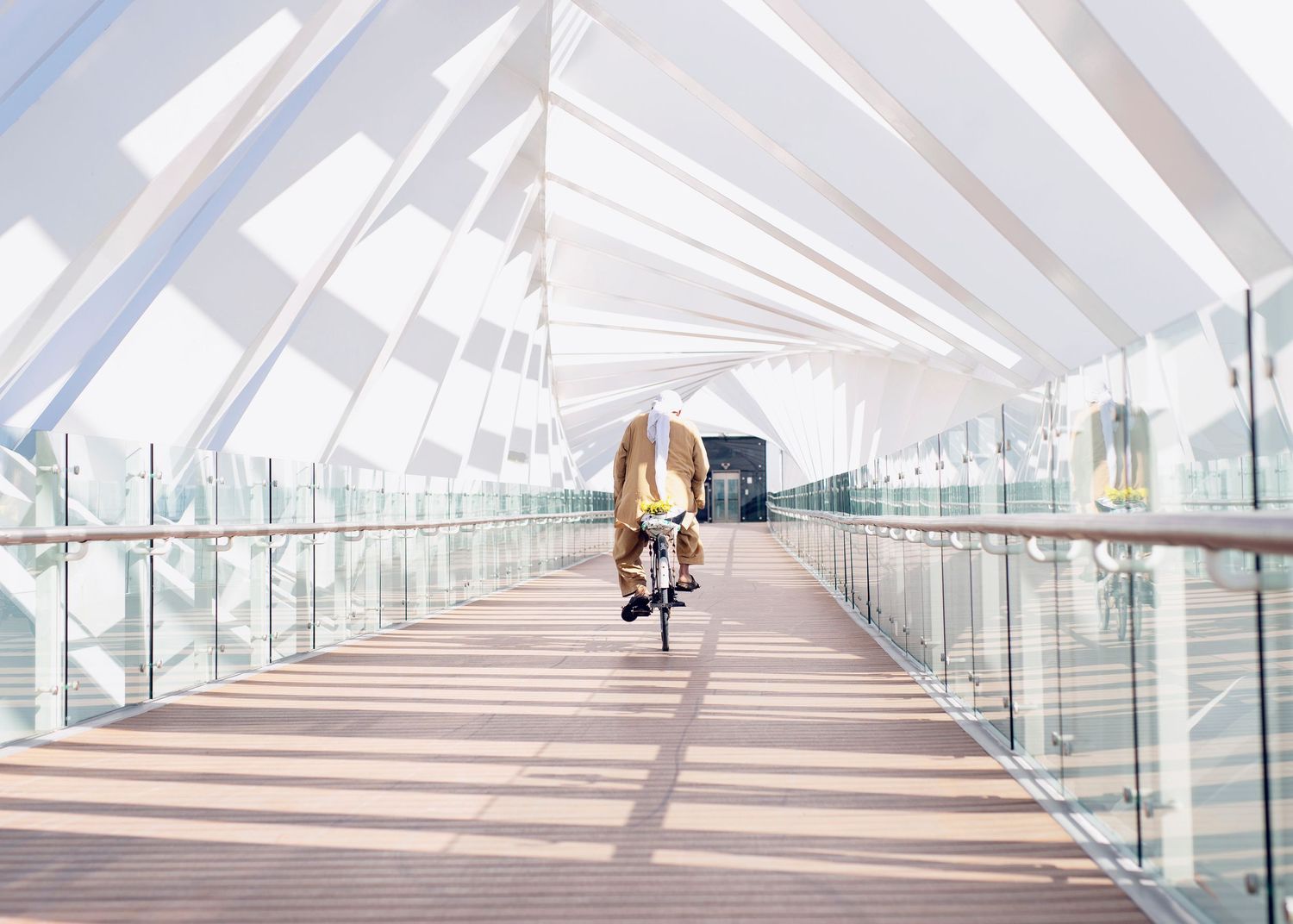 Person on bicycle rides through modern glass-covered pedestrian bridge with geometric white beams in bright daylight.