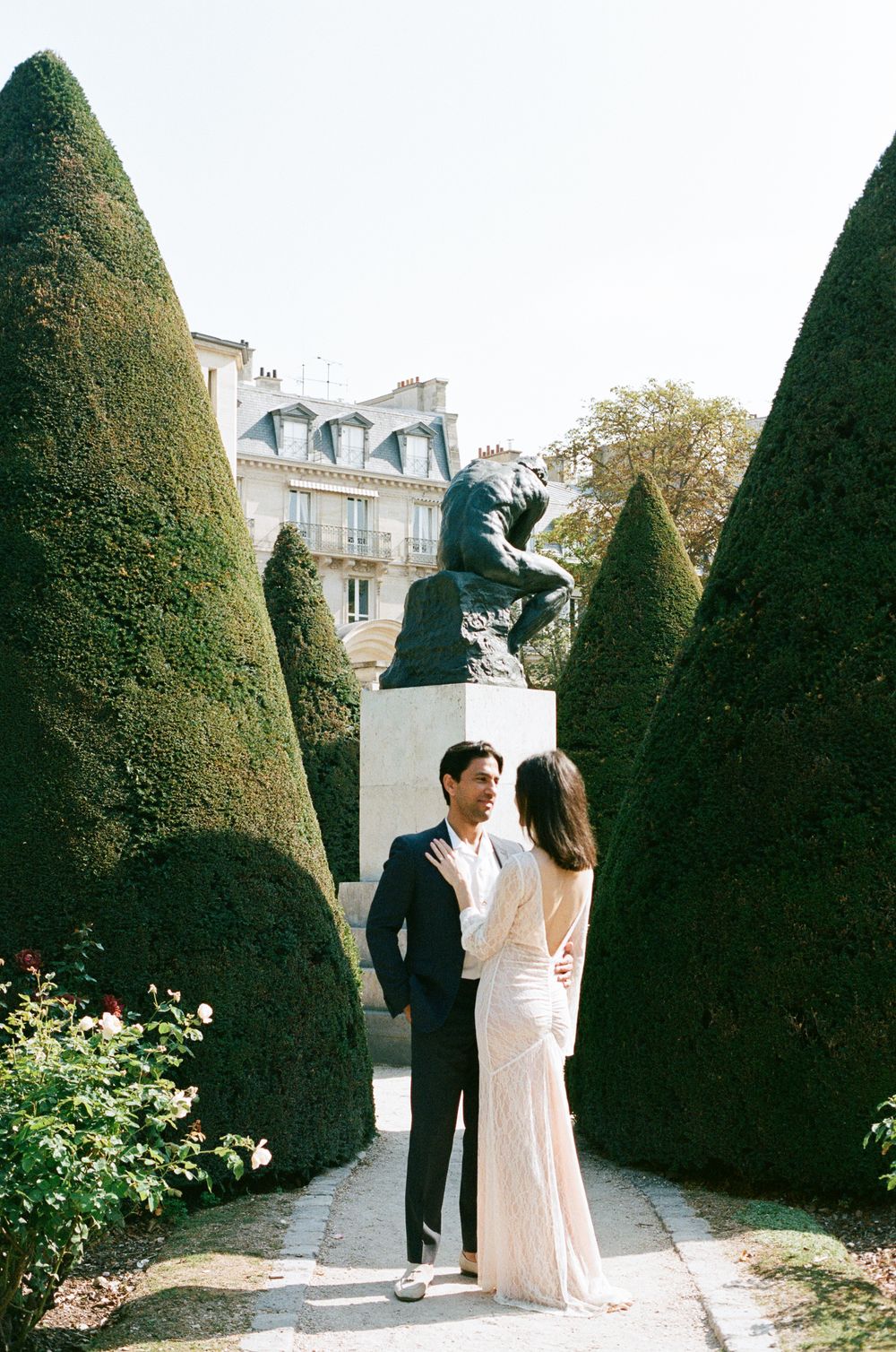 Couple sharing a romantic moment in a Parisian garden with sculpted hedges and a bronze statue backdrop.