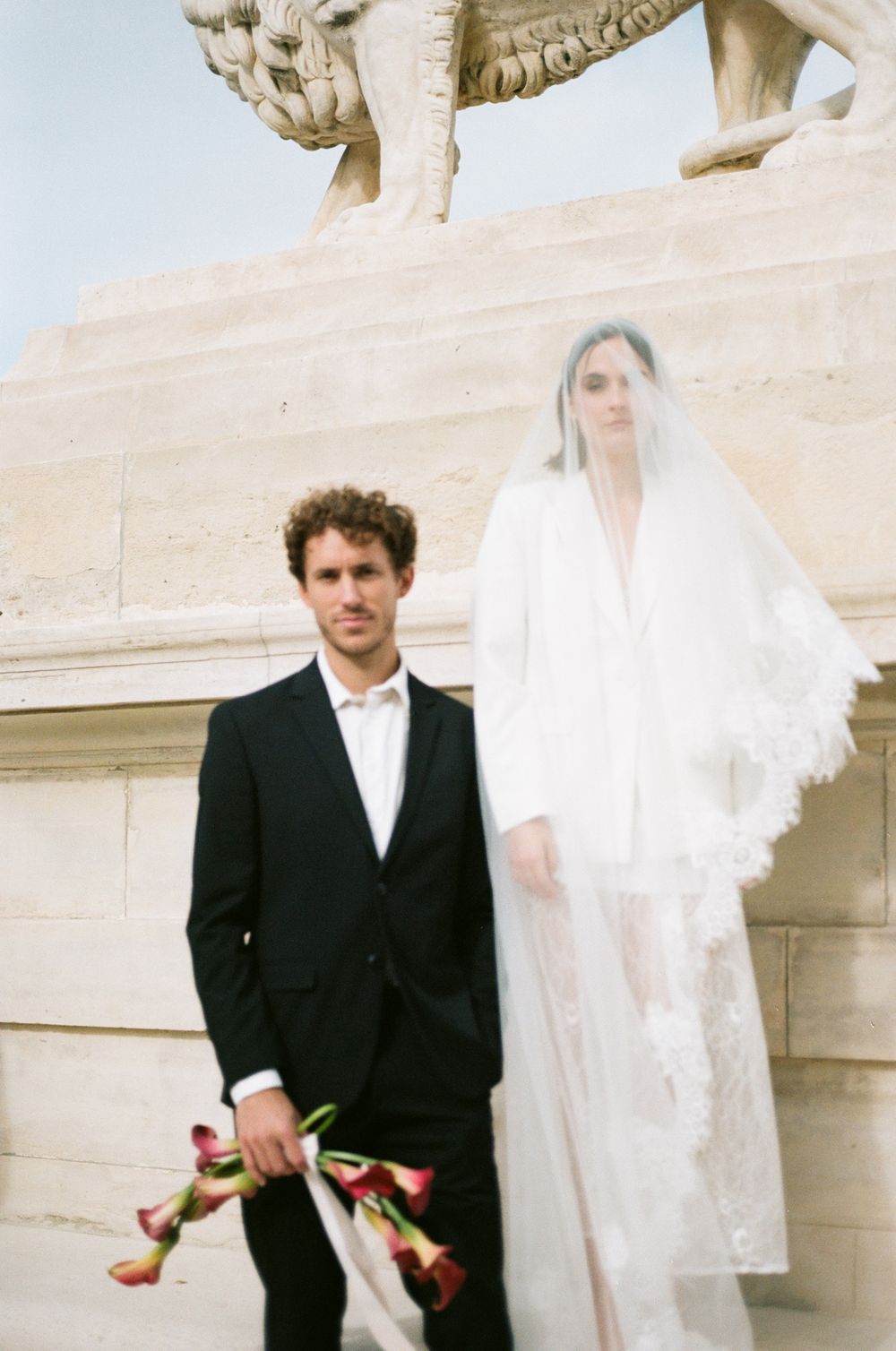 A groom in a black suit holds red calla lilies beside a bride in a white gown and veil near a stone lion statue.