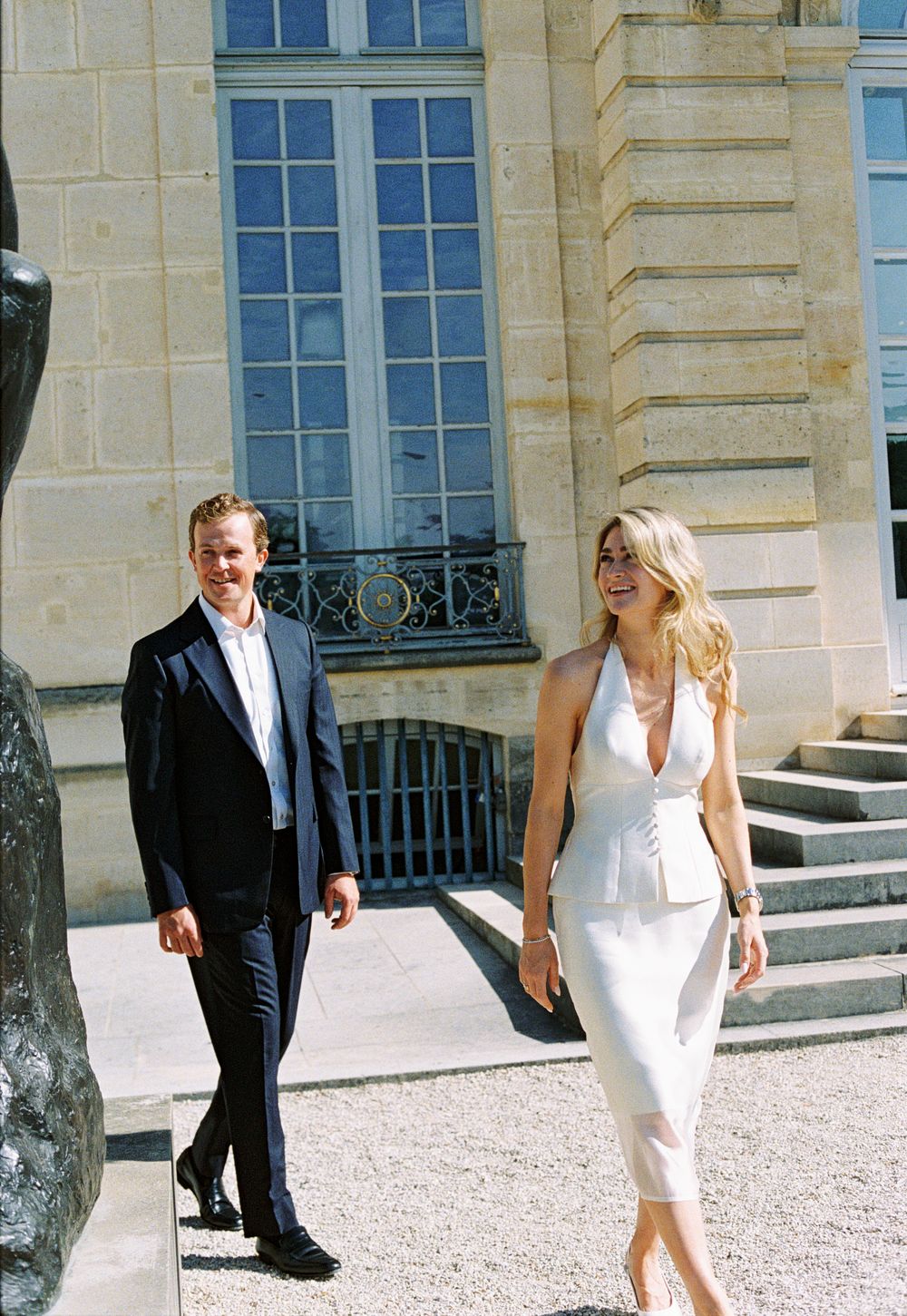 A man in a dark suit and woman in a white halter dress walk outside a classic European building.