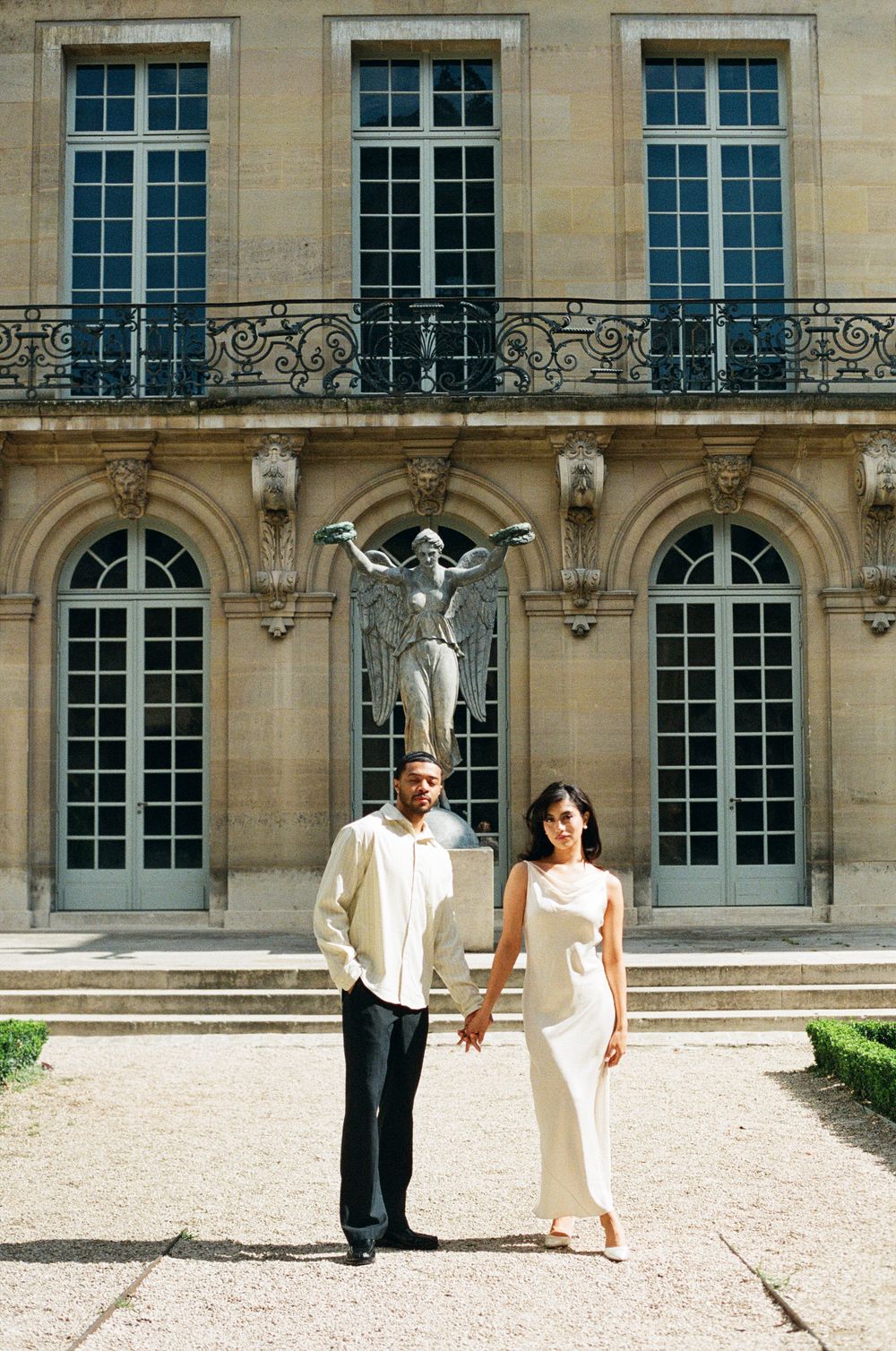 Couple in elegant attire holding hands before a classical Parisian building with a winged statue centerpiece.