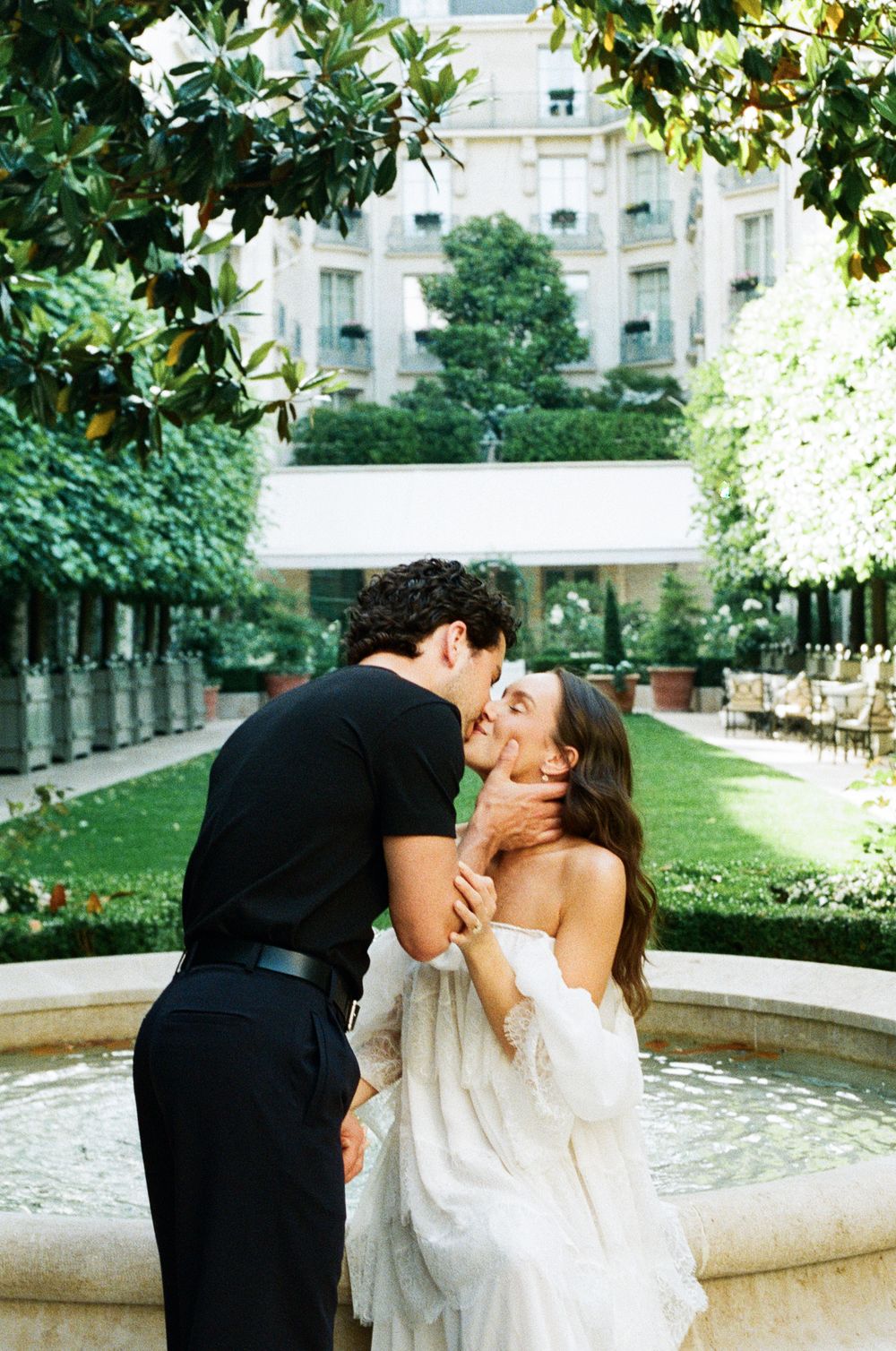 Couple sharing a romantic kiss by a fountain in an elegant garden courtyard, bride in white off-shoulder gown.