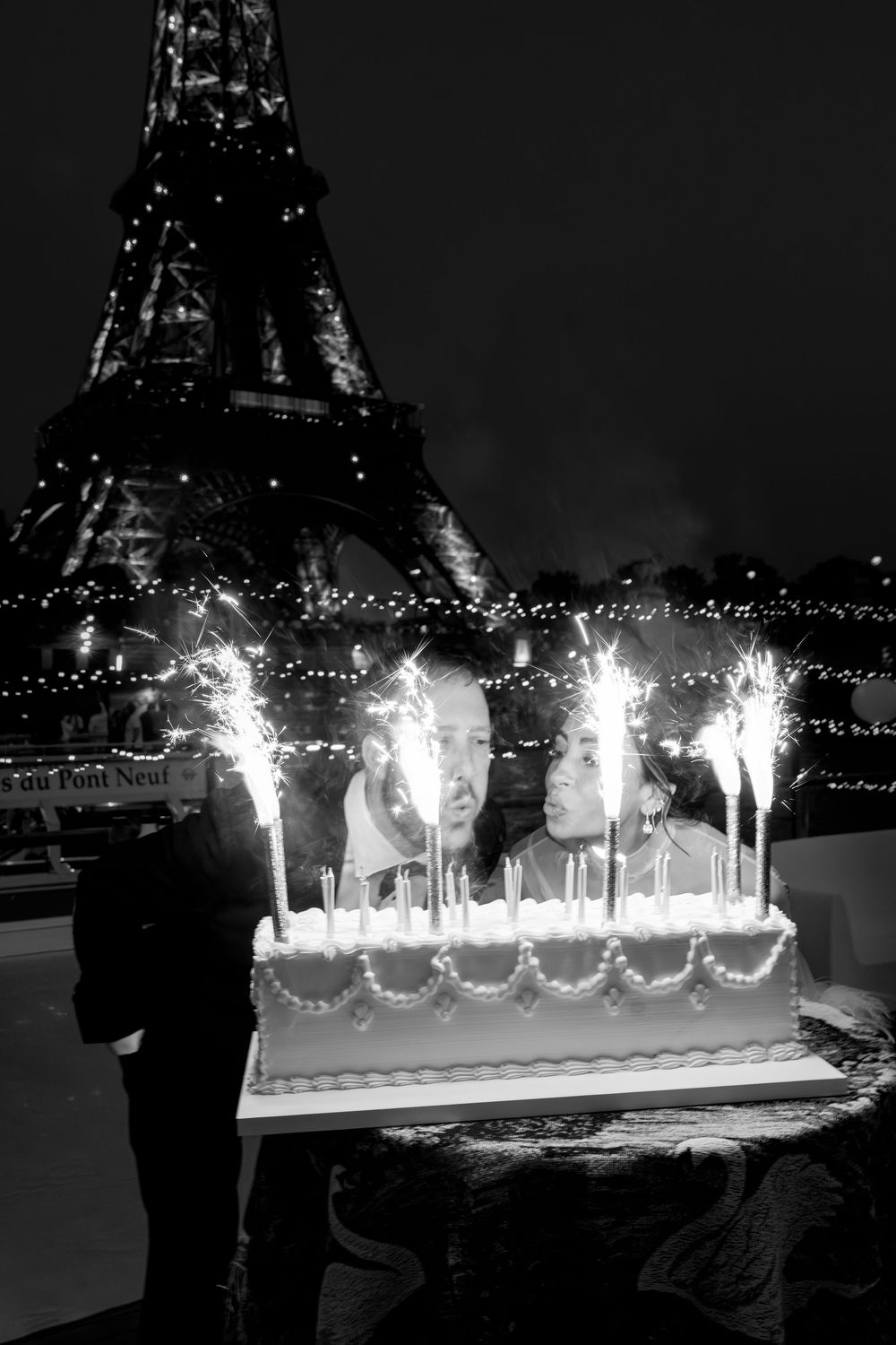 Black and white photo of two people with a sparkler birthday cake in front of the Eiffel Tower at night in Paris.