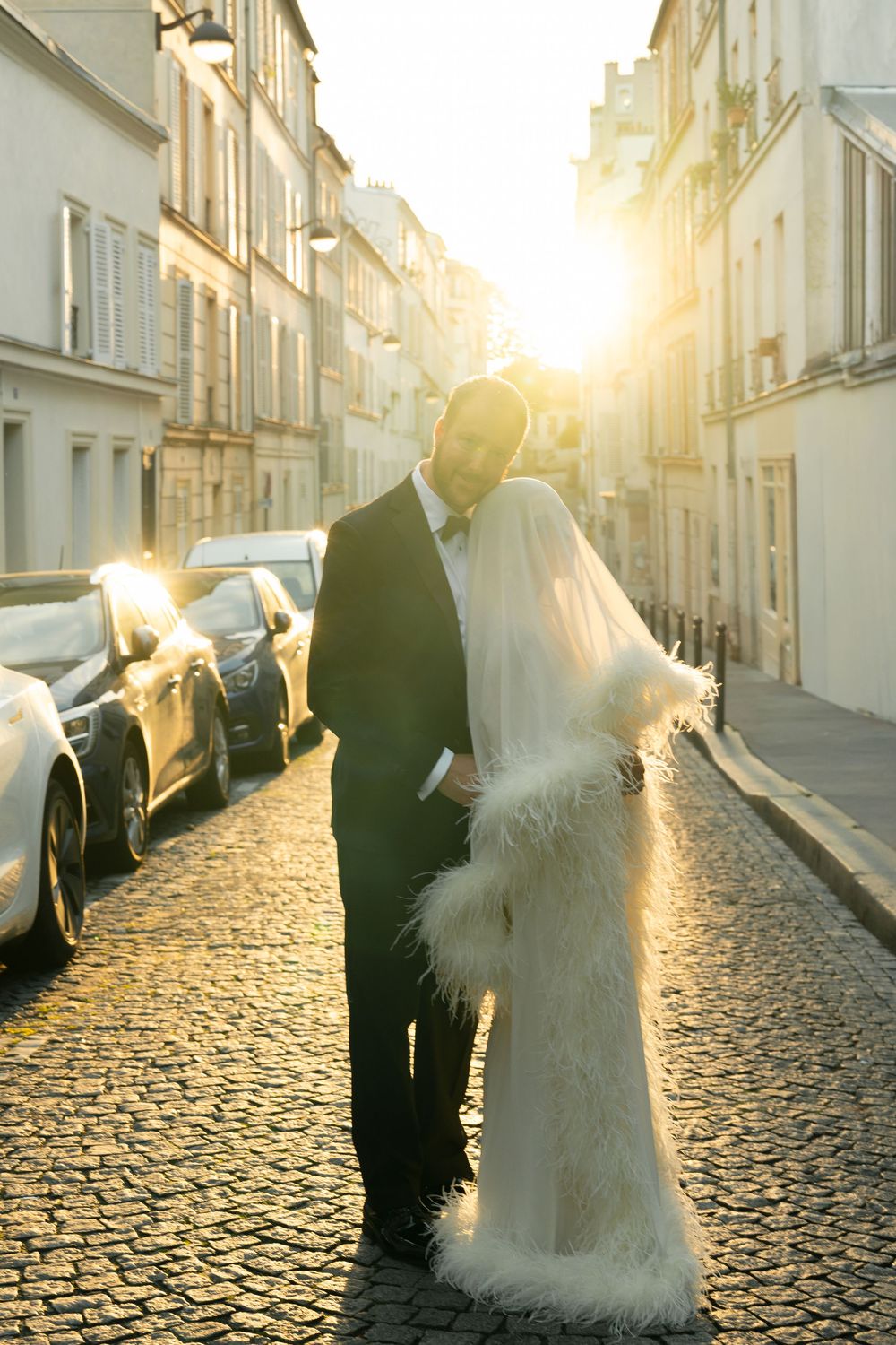 Bride and groom kissing on cobblestone Paris street at golden sunset, bride wearing white fur-trimmed cape veil.