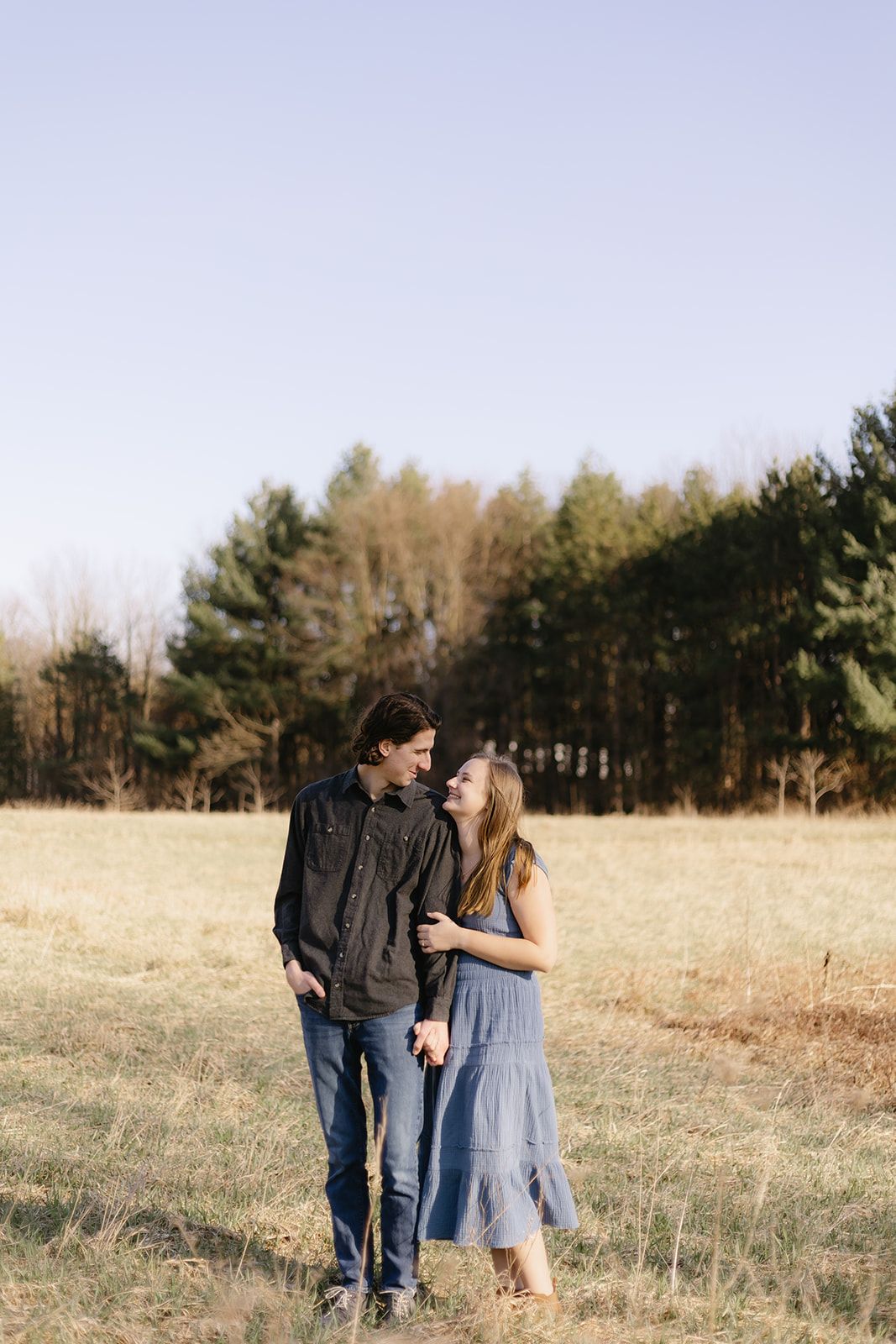 A couple walks together in a scenic field with evergreen trees in the background.