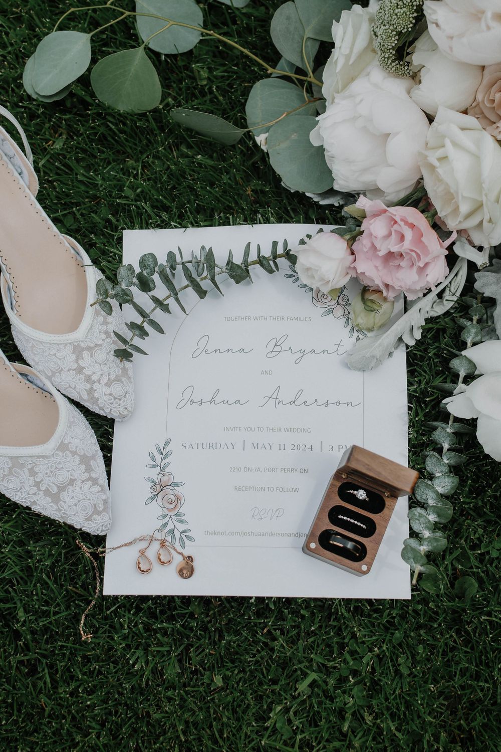 Wedding invitation with delicate floral design laid on grass alongside white lace shoes and roses.