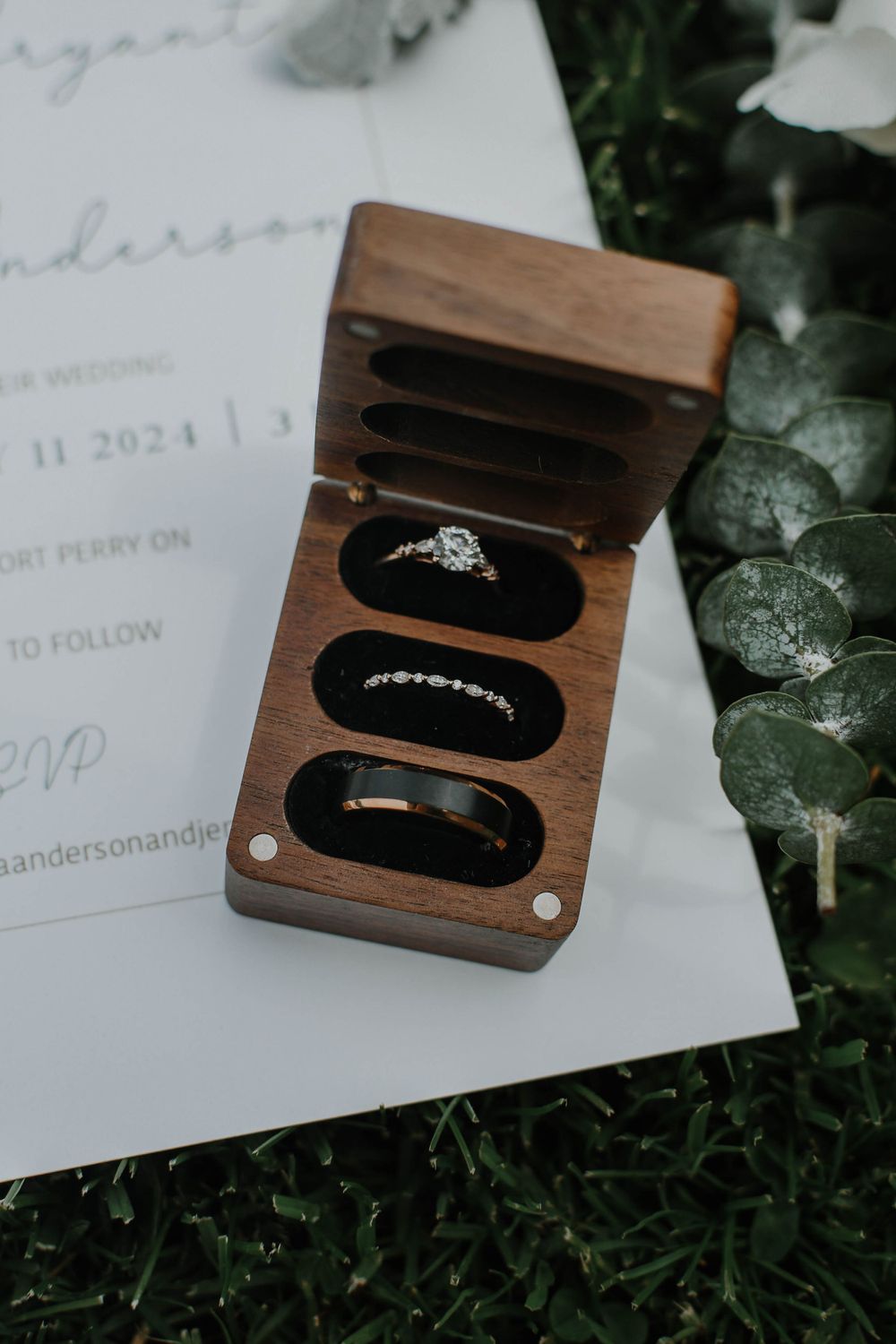 Wedding or engagement rings displayed in a wooden ring box holder with black velvet inserts on grass next to paper.