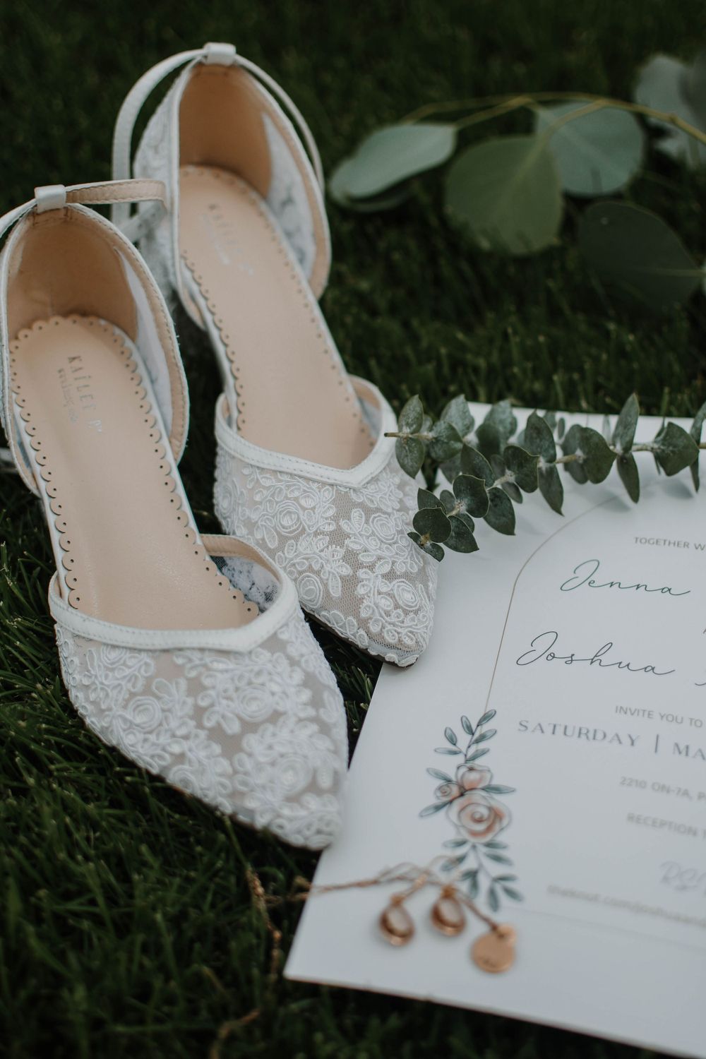 White lace wedding shoes and floral invitation card arranged on grass with eucalyptus leaves in soft lighting.