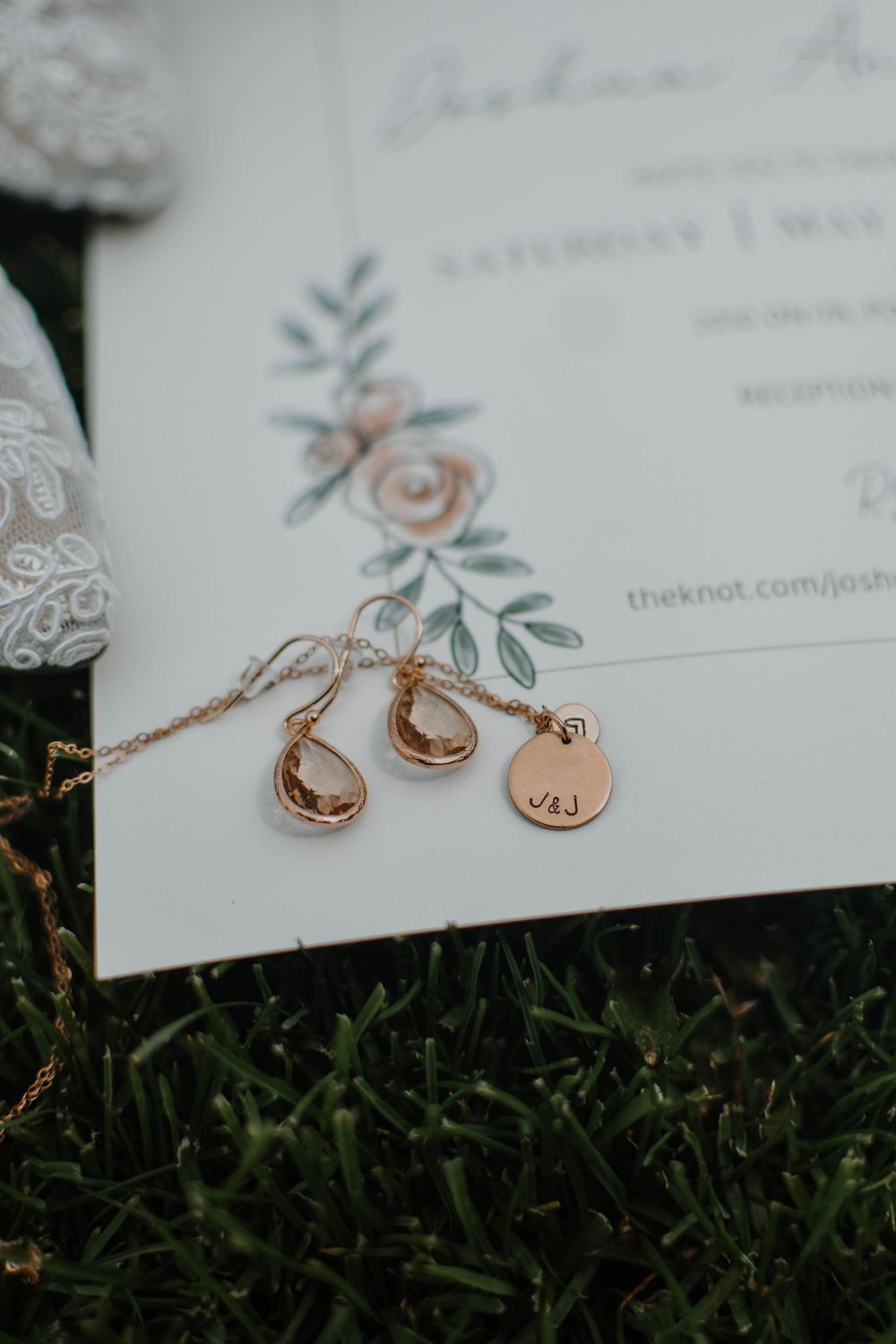 Rose gold wedding jewelry and invitation details with floral design resting on green grass.