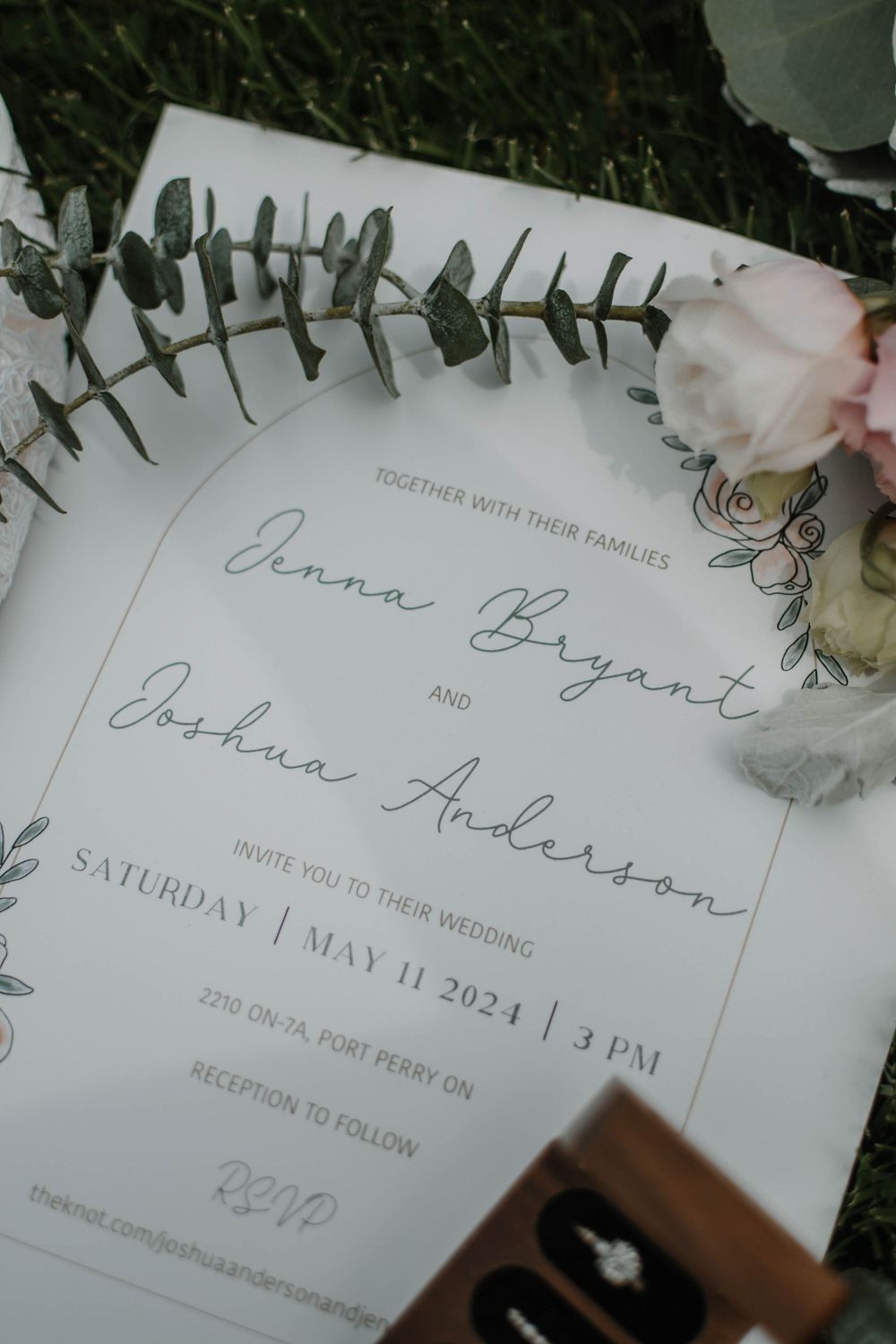 Elegant wedding invitation layout with eucalyptus leaves, pink roses and a ring box visible in romantic natural lighting.