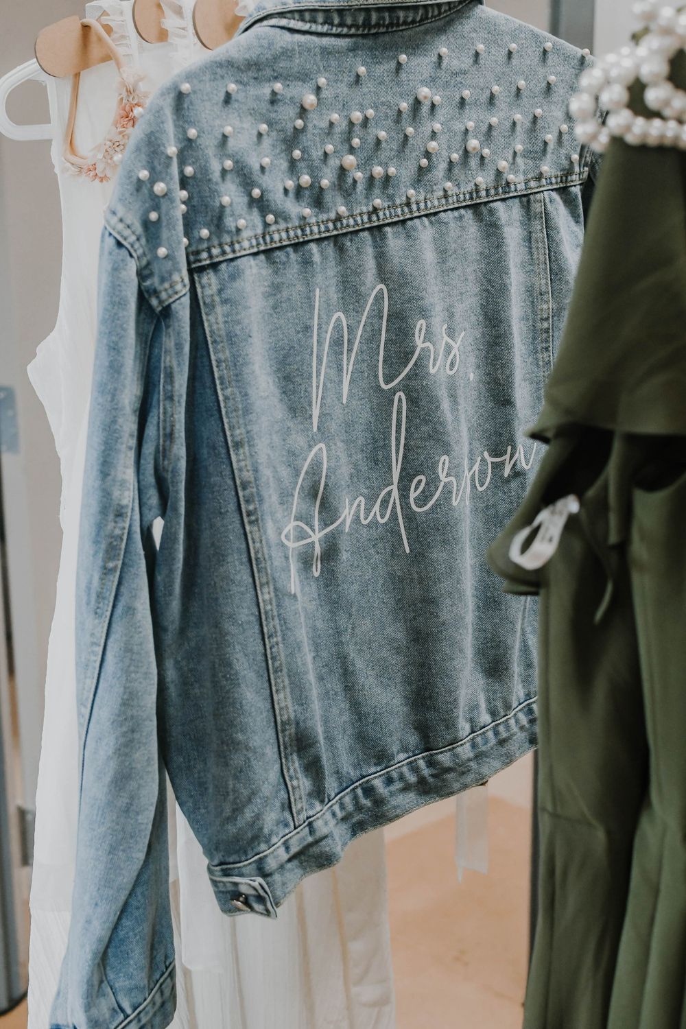 A light wash denim jacket with pearl embellishments and 'Mrs.' embroidery hanging on display.