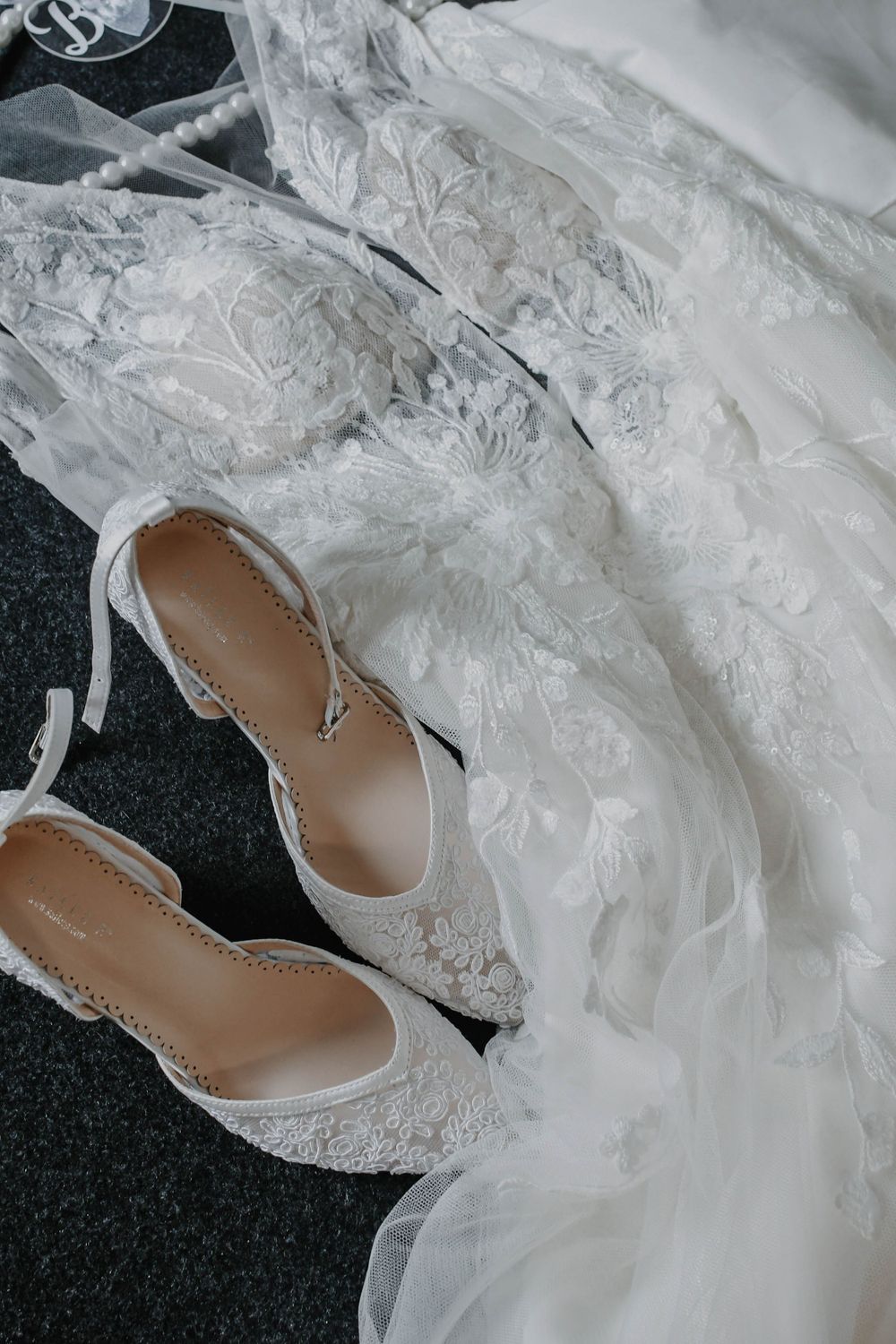 White lace wedding shoes and delicate tulle fabric laid out on dark carpet, creating an elegant bridal styling detail.
