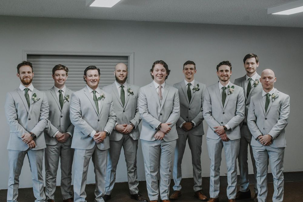 A group of eight groomsmen wearing matching light gray suits and ties pose together in a row against a gray wall.