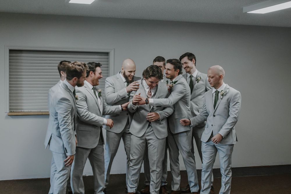 A group of groomsmen in light grey suits huddle together looking at something on a phone in an indoor setting.