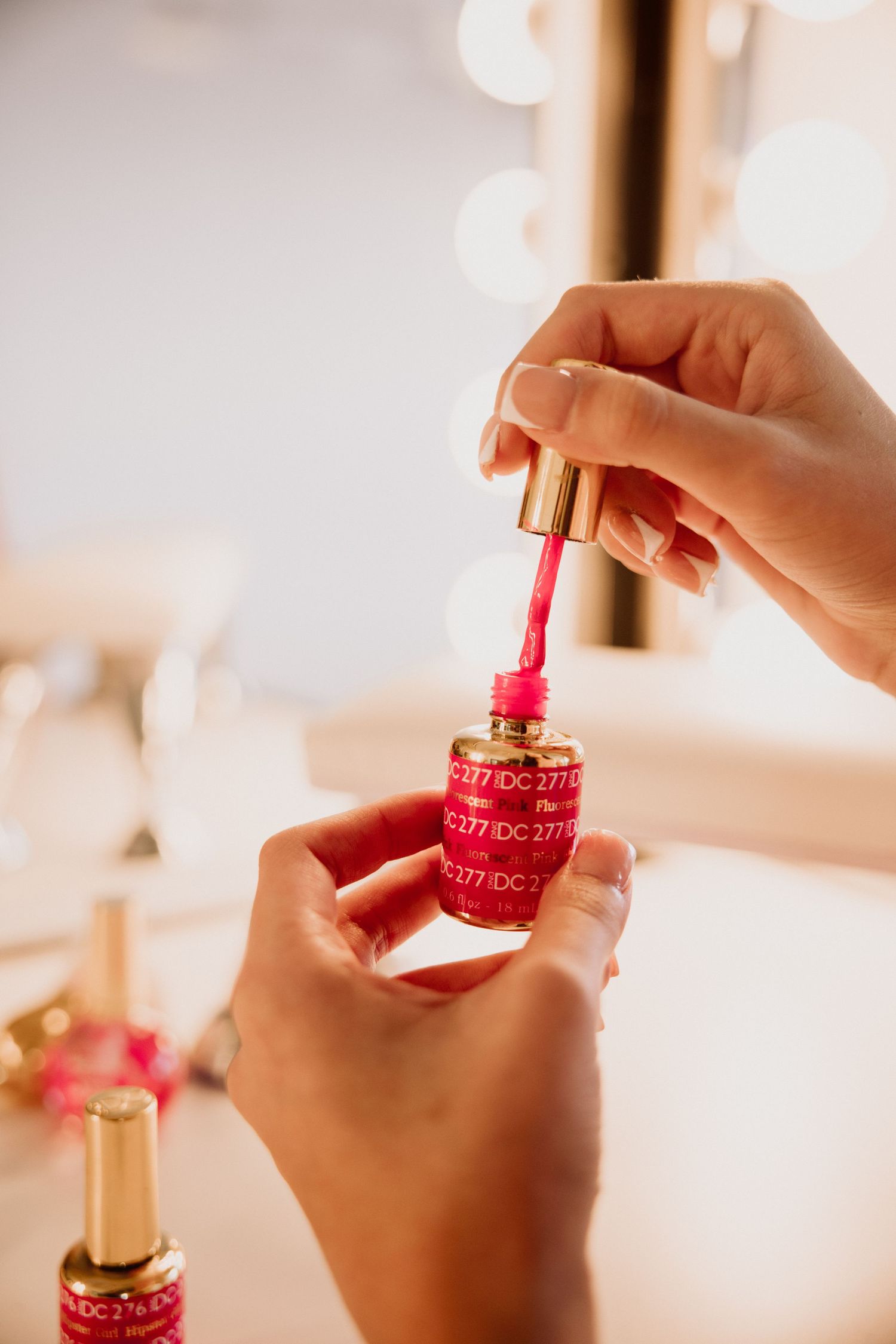 Hand applying bright pink nail polish from a luxurious gold-capped bottle in warm lighting.