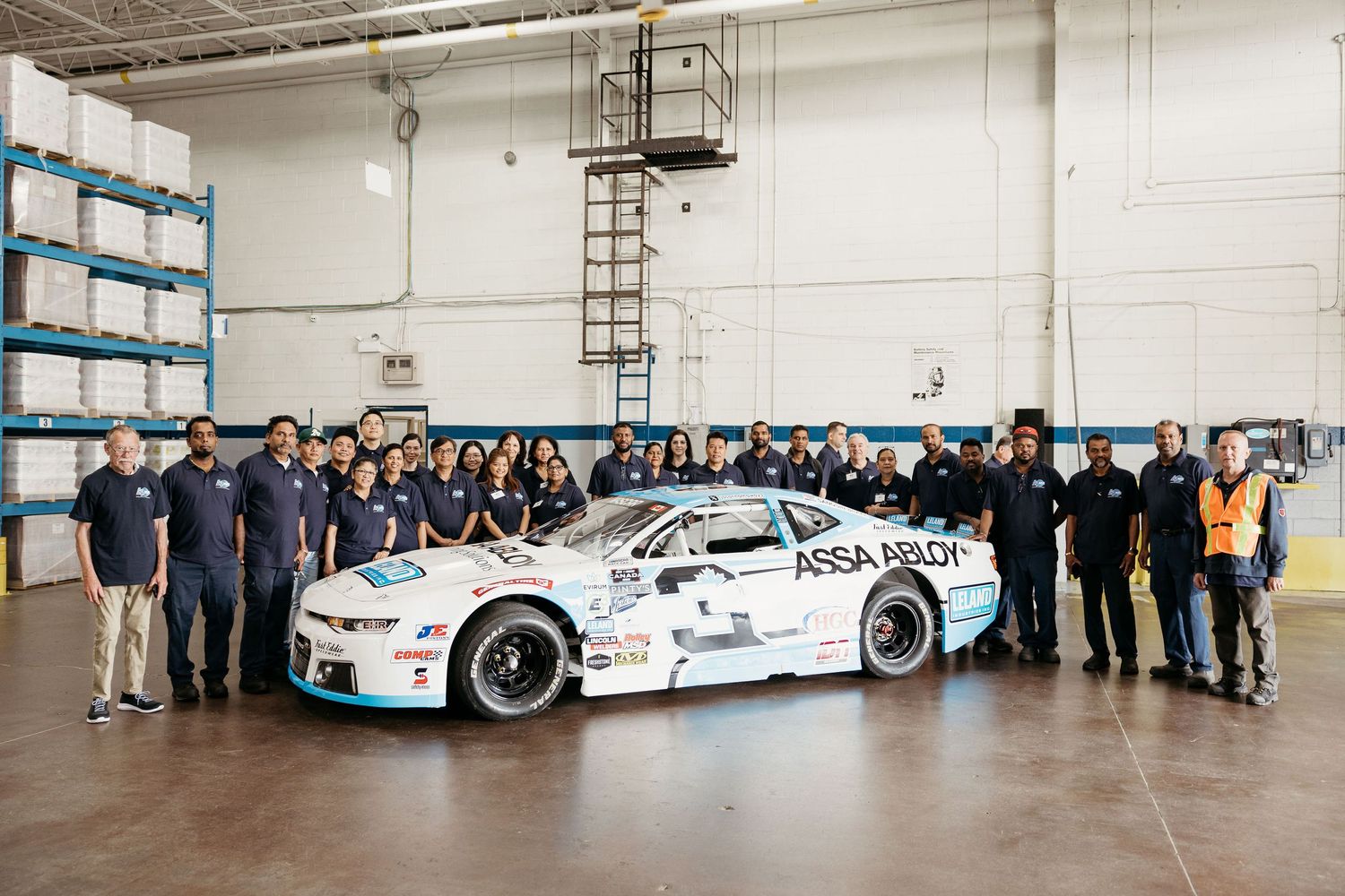 Wide panoramic view of employees gathered in warehouse with race cars and delivery vehicles.