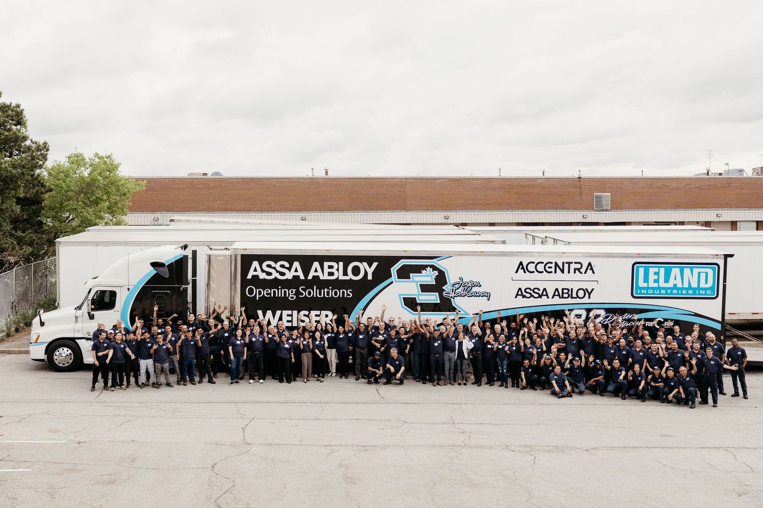 Panoramic group photo of employees standing in front of warehouse building and delivery trucks.