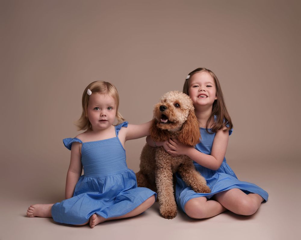 Two young girls in matching blue dresses pose with goldendoodle dog against neutral backdrop.