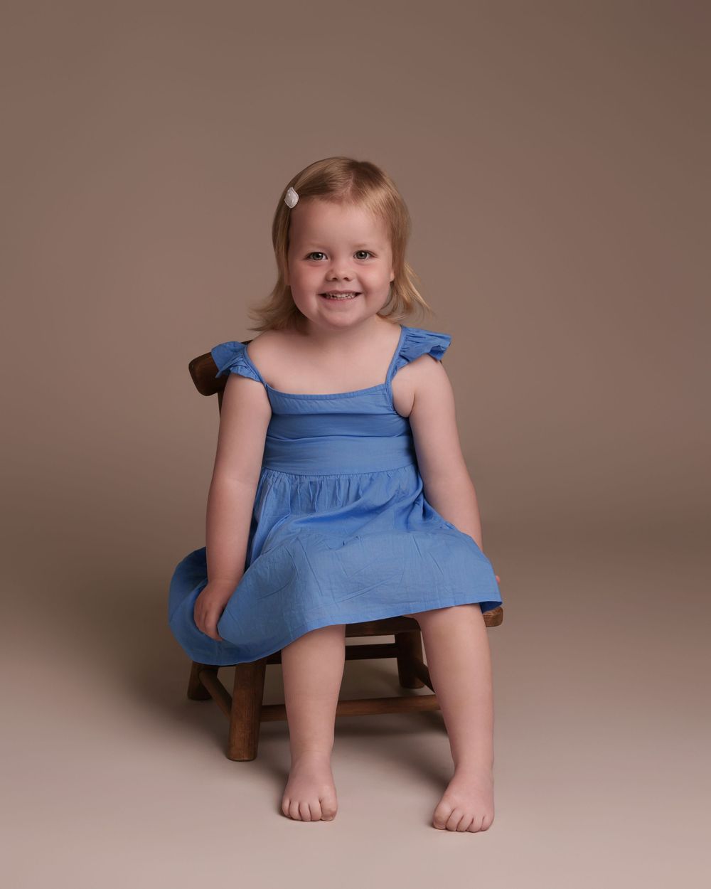 Young girl in blue summer dress sits on wooden stool against neutral backdrop.