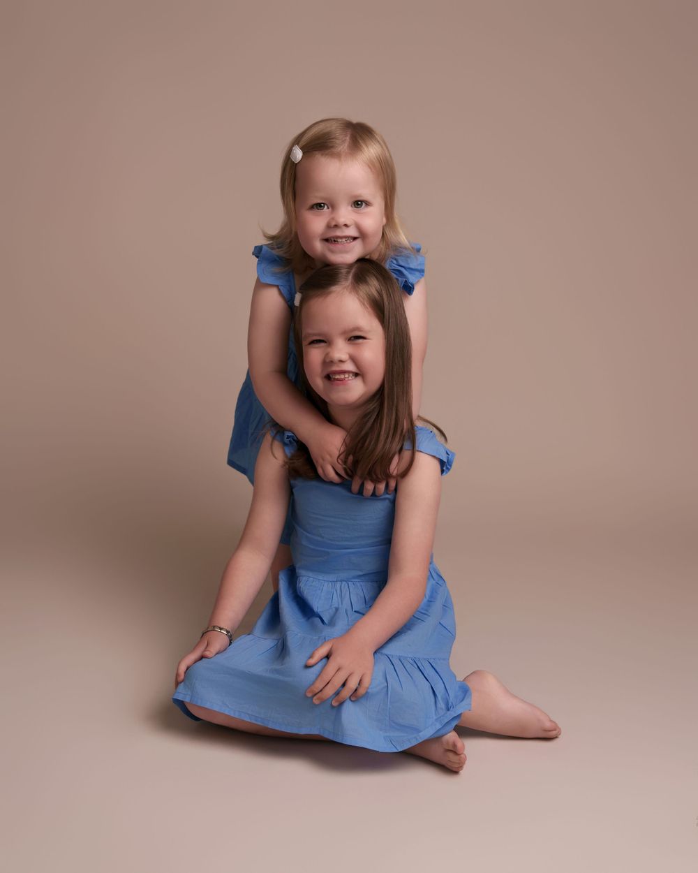 Two sisters in matching blue dresses pose together against neutral studio backdrop.