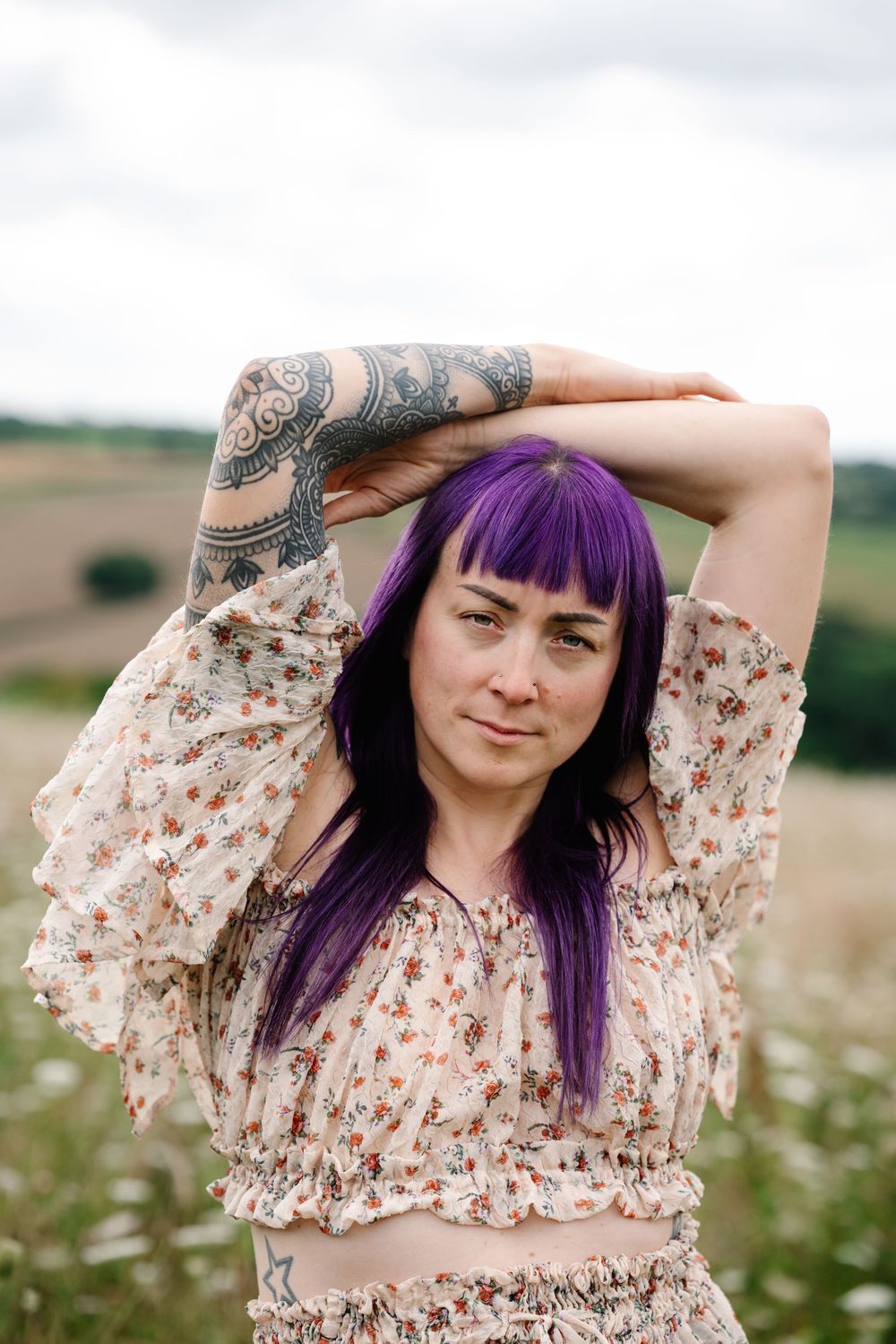 Woman with vibrant purple hair poses playfully in a floral dress against a rural landscape with wildflowers.