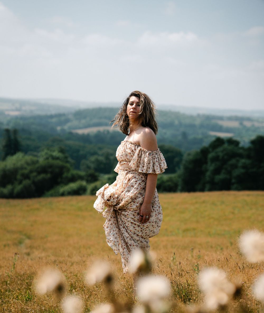 A panoramic view of a person in a floral dress standing in a golden meadow with mountains in the background.
