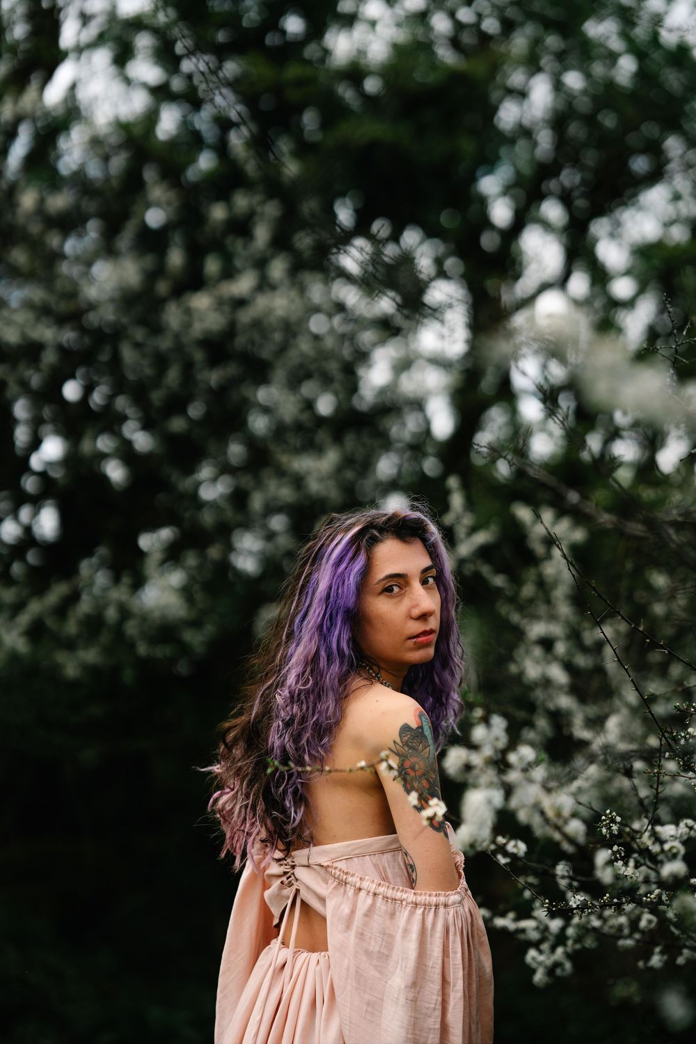 Person wearing pink dress with purple hair poses among blooming spring branches.