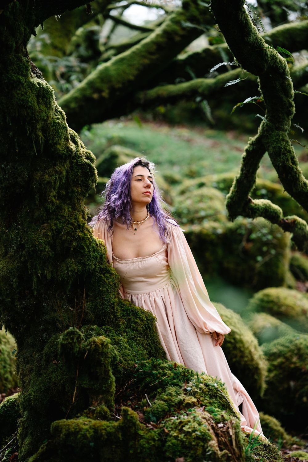 Person in pink dress poses against moss-covered tree in enchanted forest setting.