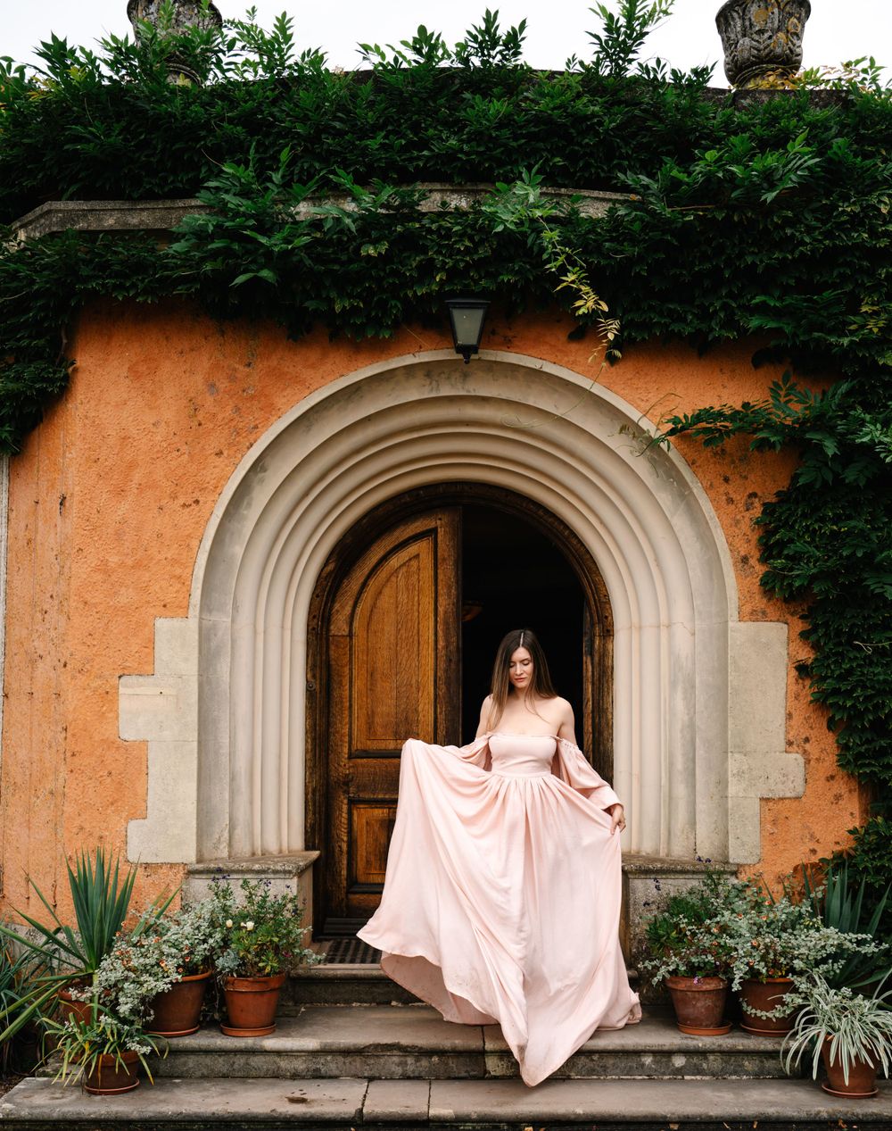A person in a flowing pink dress poses in front of an arched doorway surrounded by ivy.