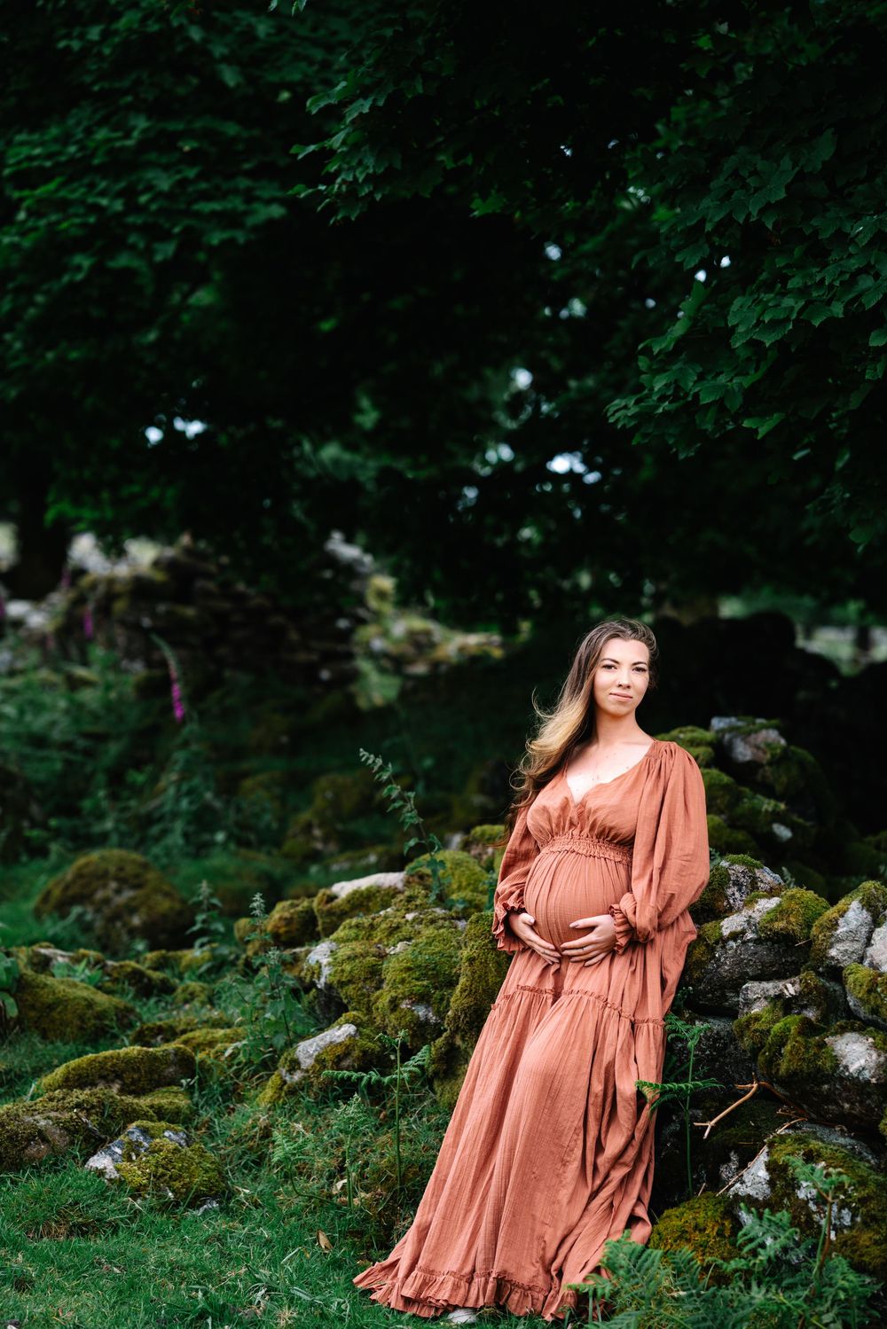 A person in a flowing terracotta dress poses gracefully against a mossy stone wall in a lush forest setting.
