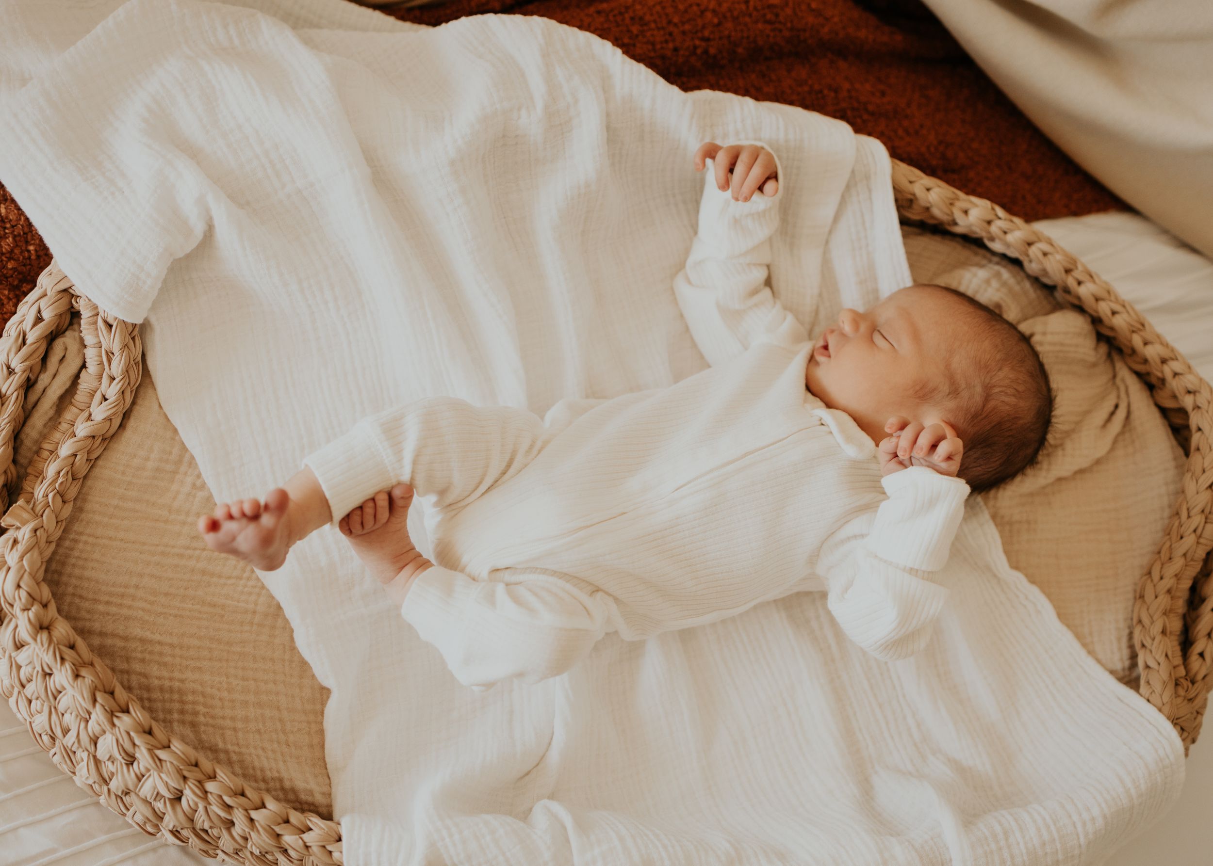 A newborn baby in a white christening gown rests peacefully in a woven bassinet with beige bedding.