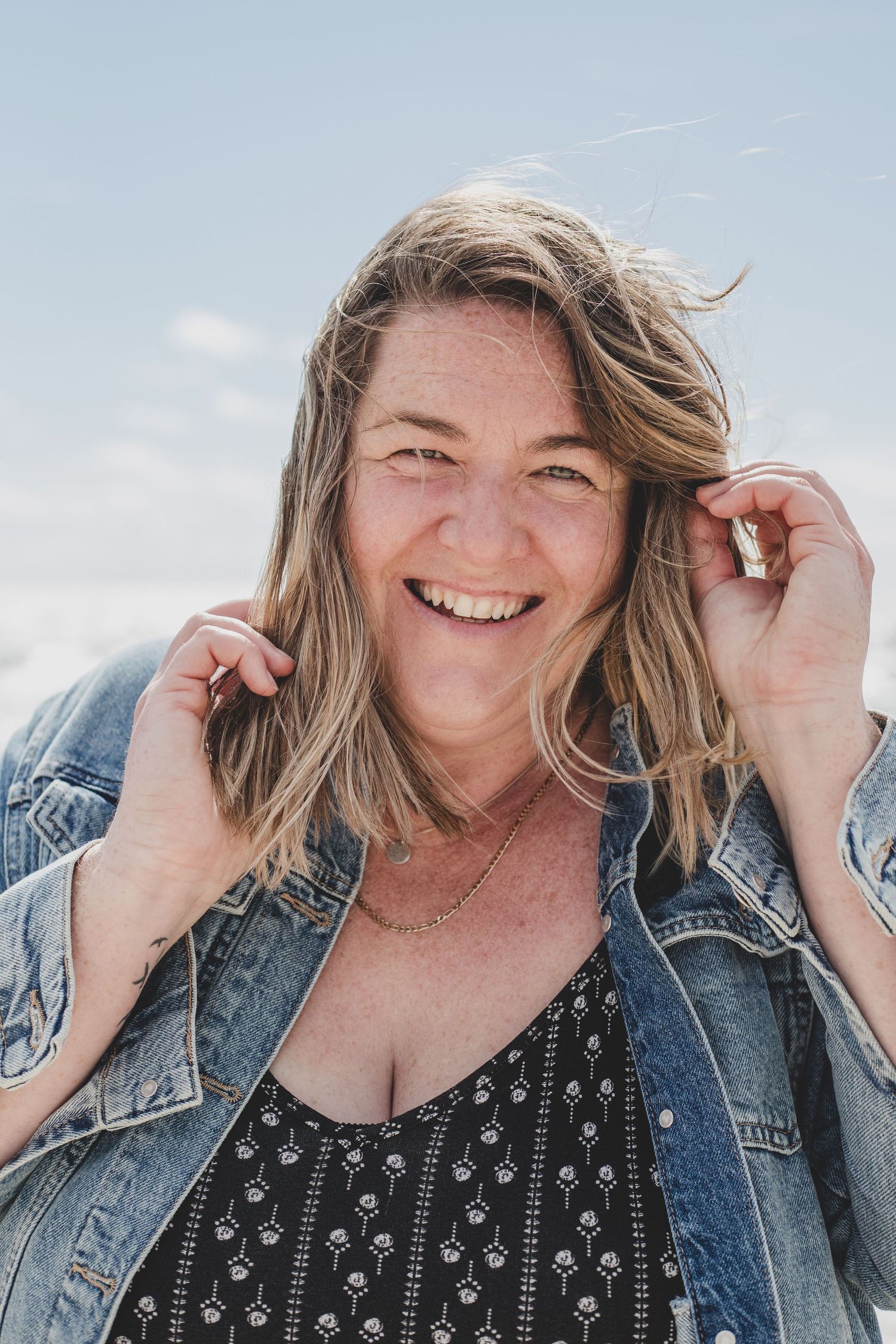 A candid portrait of a woman with windswept hair wearing denim jacket against blue sky.