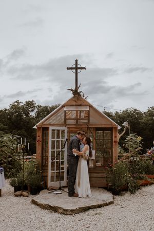 A couple embraces outside a charming wooden greenhouse structure with cross details.