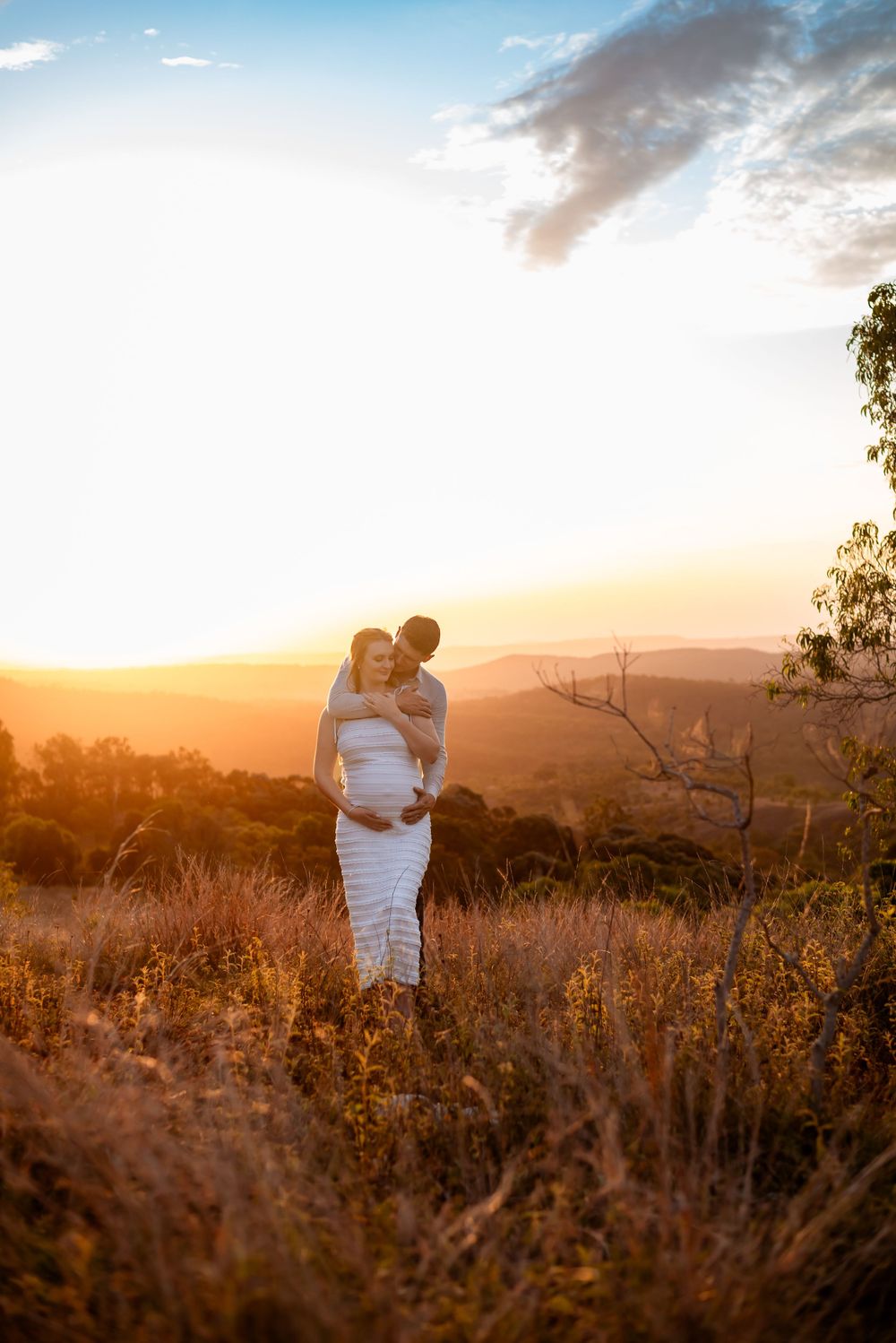 Couple embraces in golden field during dramatic sunset with mountain backdrop.